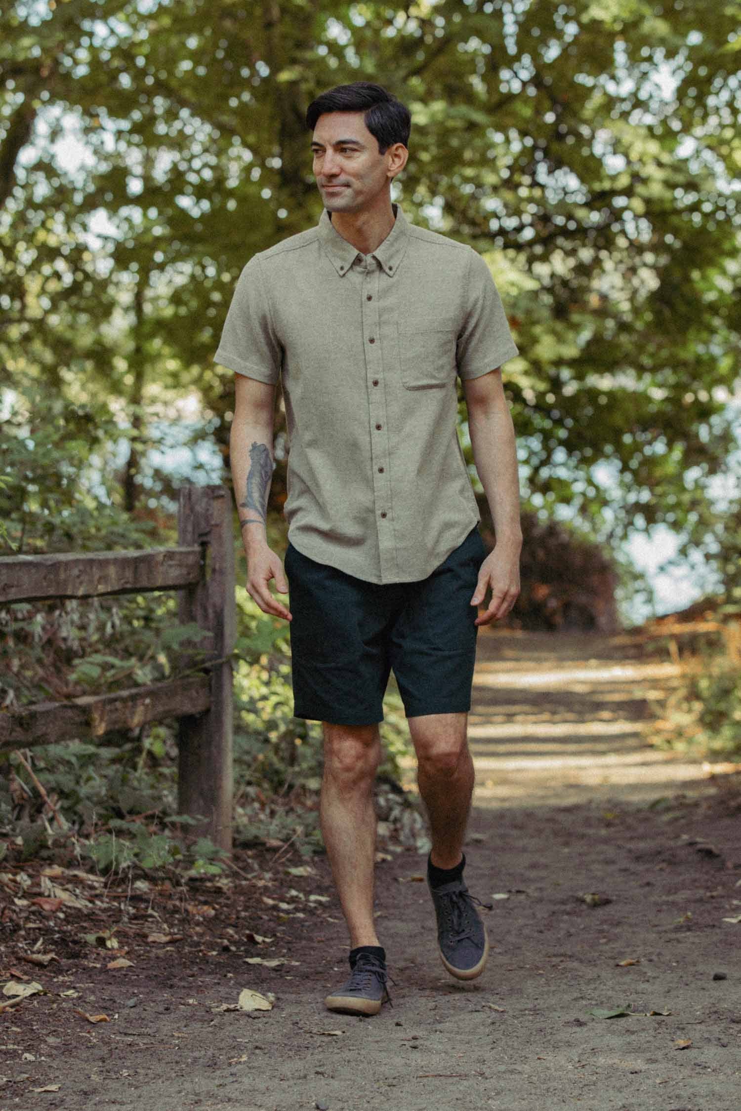 A man wearing the Bridge & Burn Bryce Shirt in Olive Twill and dark shorts walks along a shaded dirt path in a wooded area, with sunlight filtering through the trees.