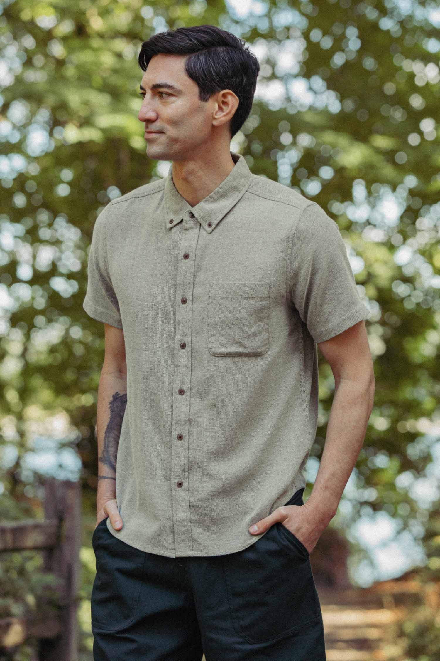 A man stands outdoors in the Bridge & Burn Bryce Shirt—an olive twill short sleeve button-down—paired with dark pants, hands in his pockets. Trees and greenery fill the background.