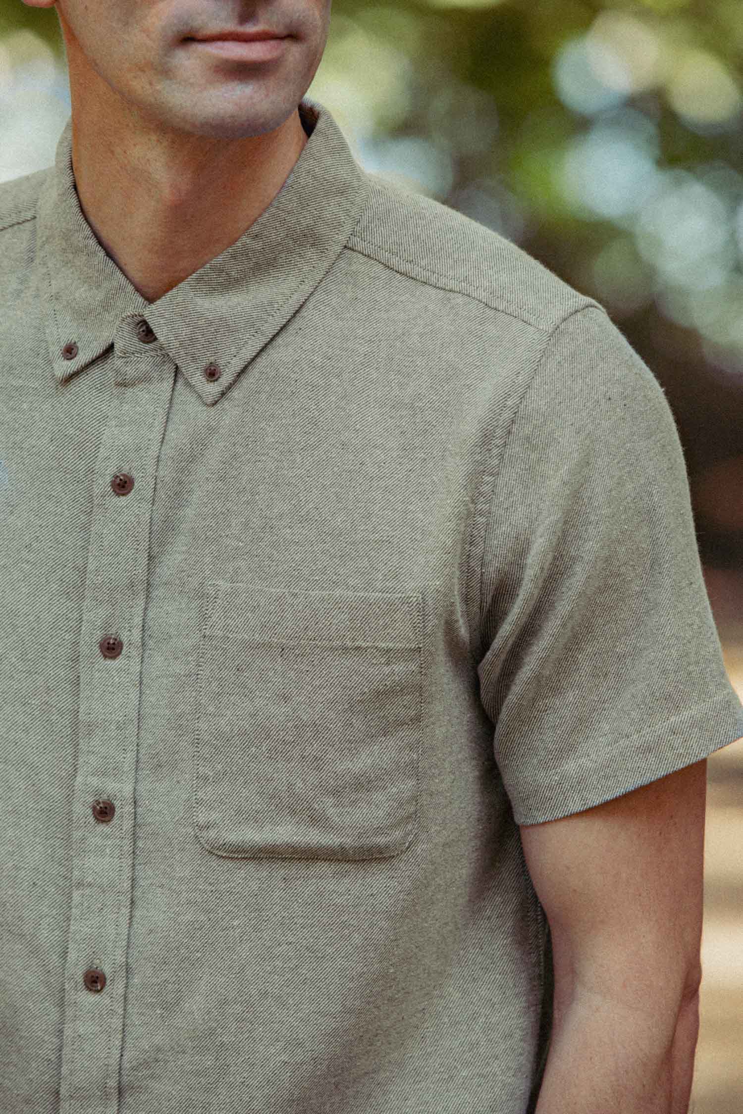 A man wears the Bridge & Burn Bryce Shirt in olive twill, a short sleeve button-down with a chest pocket, while standing outdoors against a blurred natural background.
