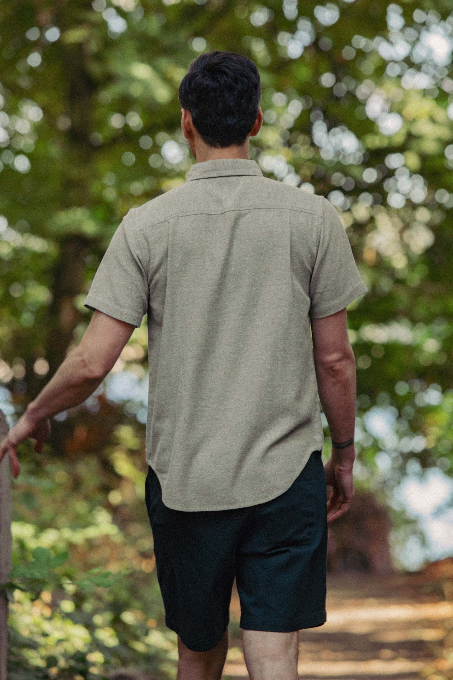 A man with short dark hair, wearing the Bridge & Burn Bryce Shirt in Olive Twill and black shorts, walks along a shaded outdoor path surrounded by green trees, viewed from behind.