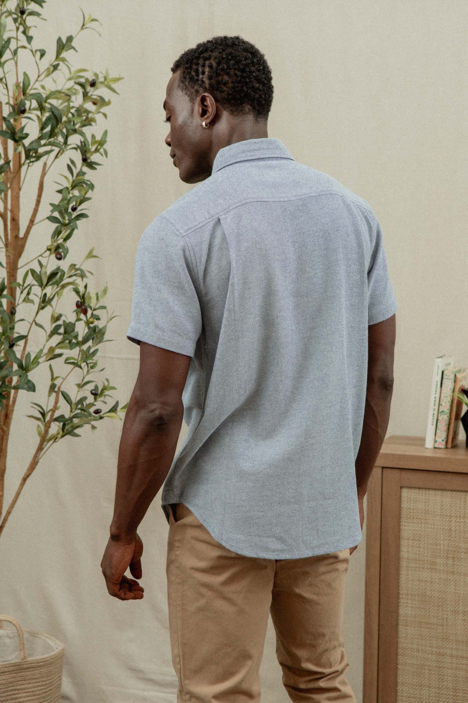 A man stands indoors facing away, wearing the Bridge & Burn Bryce Shirt in Denim Twill and tan pants. Behind him are a leafy plant, a wooden cabinet with books, and decor in neutral tones.