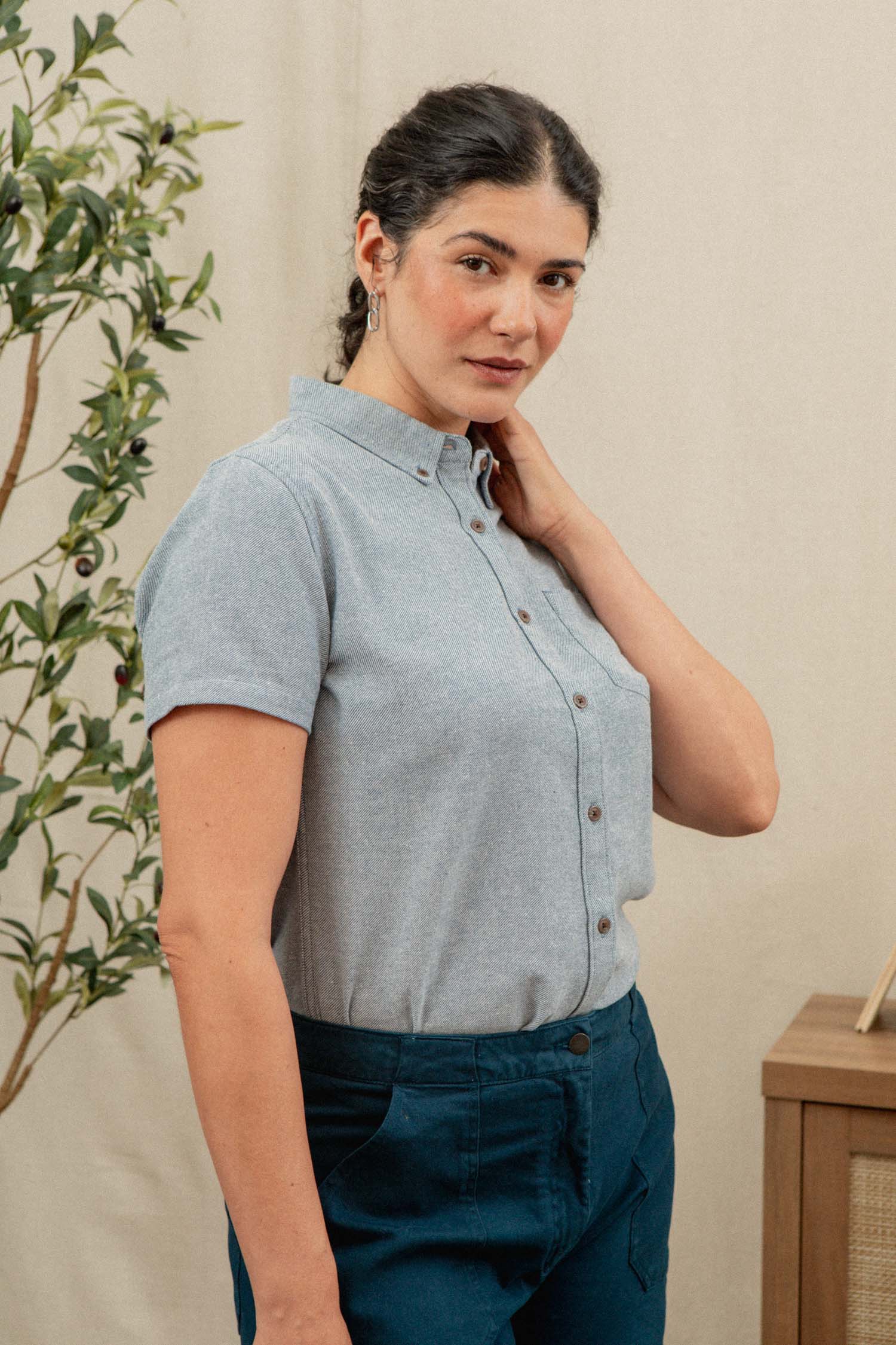 A woman with dark braided hair wears the Bridge & Burn Juniper Shirt in Denim Twill— a relaxed fit, short sleeve button-down made from recycled cotton—paired with blue pants, standing indoors by a plant and wooden furniture.