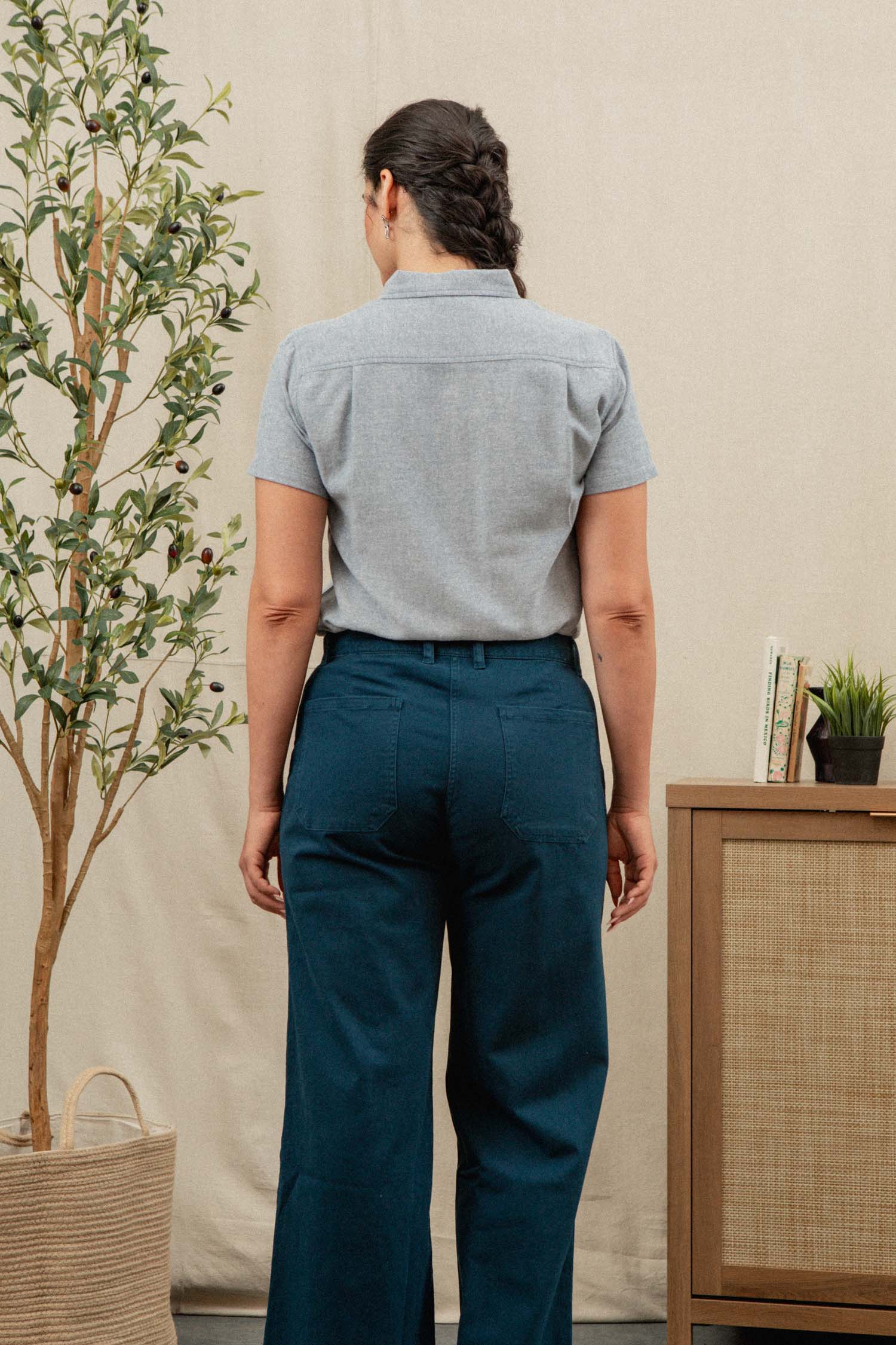 A woman stands indoors with her back to the camera, wearing the Bridge & Burn Juniper Shirt in Denim Twill. She is near a leafy potted plant and a cabinet topped with books and a small plant.