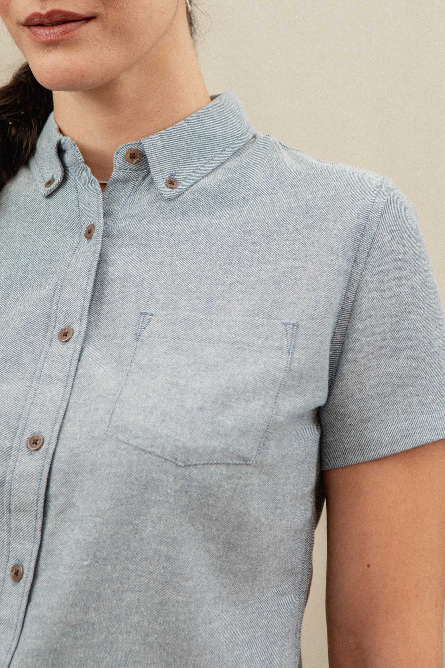 A woman with long dark hair in a ponytail wears the Bridge & Burn Juniper Shirt in Denim Twill, a relaxed fit, light blue short sleeve button-down with a chest pocket. Only her lower face, shoulders, and upper torso are visible against a neutral background.