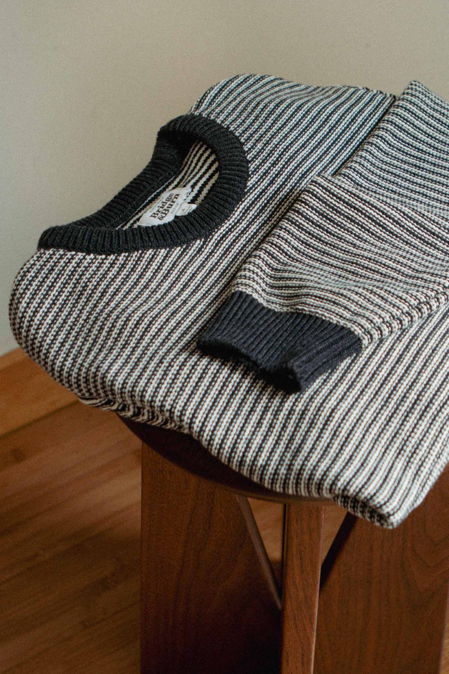 The Ansel Sweater in Charcoal Stripe by Bridge & Burn, a sustainable mid-weight sweater with black and white stripes, ribbed collar and cuffs, is crafted from recycled fibers and sits neatly on a wooden stool against a light wall.