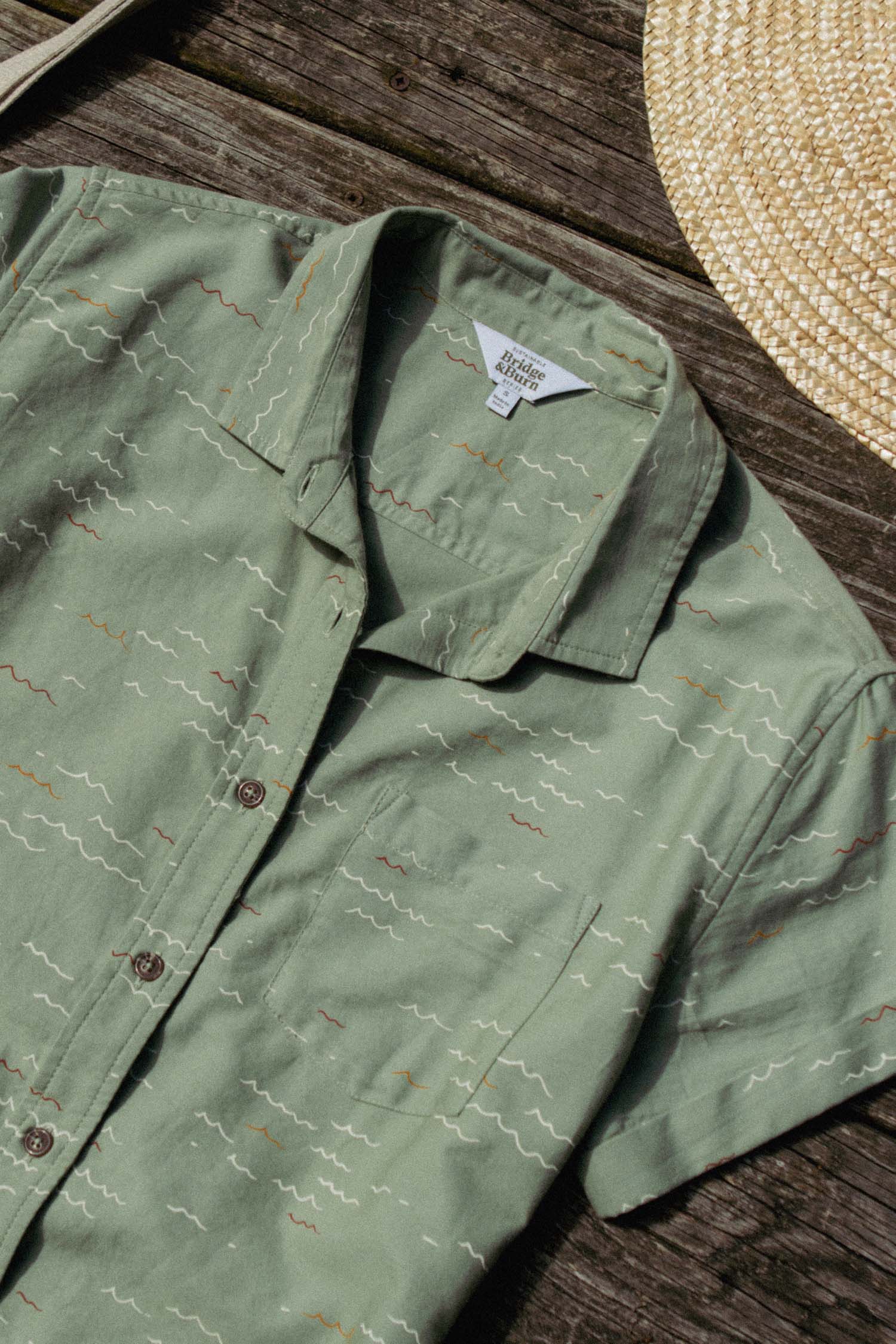 The Bridge & Burn Bea Shirt in Teal Tide Waves, an organic cotton short-sleeve button-up with a wavy pattern, is displayed on wood next to a woven straw hat.