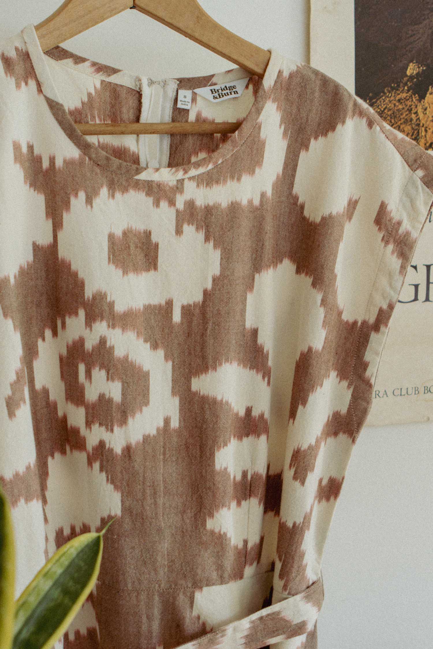 The Eloise Dress / Pop Leaf by Bridge & Burn features a beige and brown abstract geometric pattern. Hanging on a wooden hanger before a white wall and vintage poster, it’s an effortless wardrobe staple for any season.