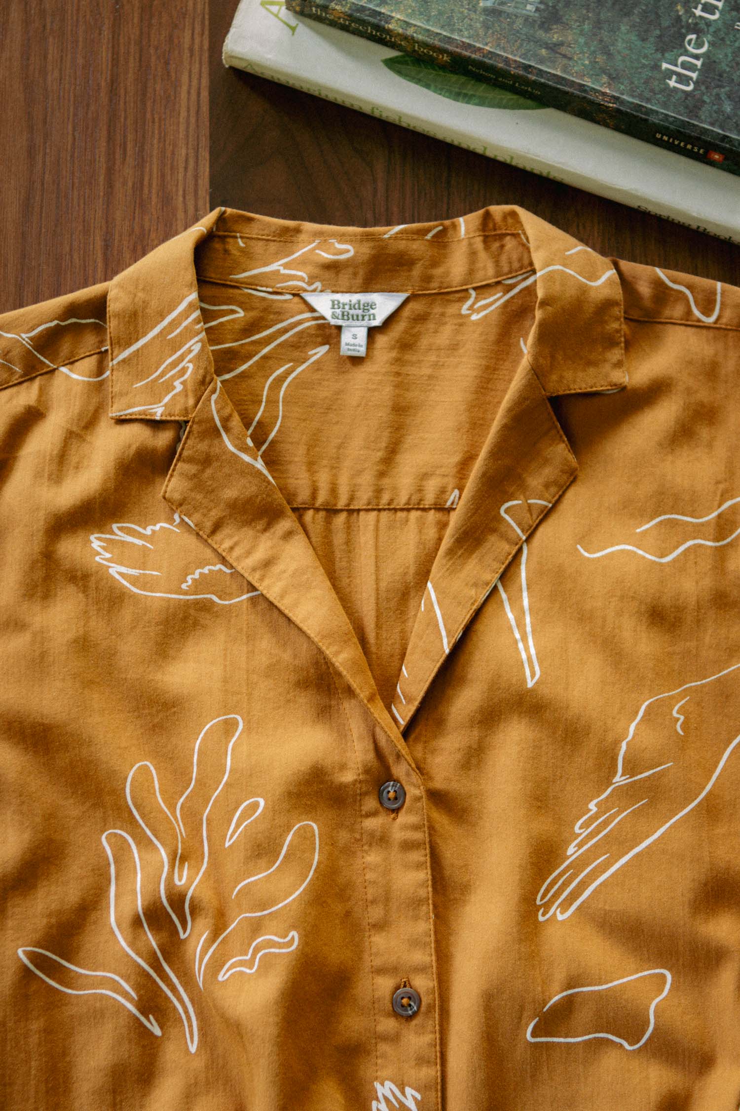 The Innes Shirt / Shoreflight by Bridge & Burn, a brown short sleeve top with white line art of abstract hands and plants, is neatly laid on a wooden surface next to two books.