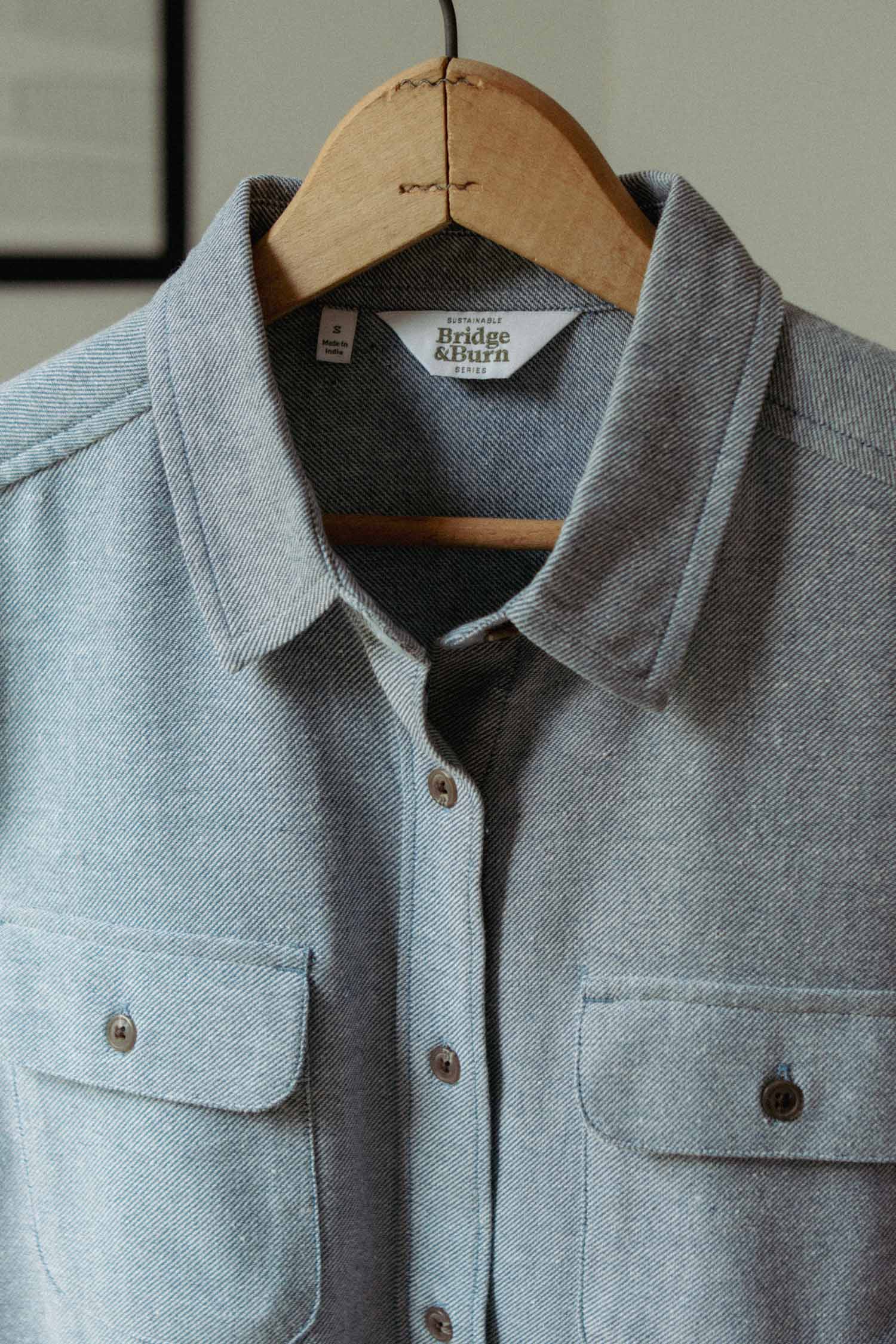 The Lenon Shirt in Denim Twill by Bridge & Burn is a light gray, recycled cotton, loose-fit button-up over-shirt with front pockets, a collared neckline, and visible stitching, shown hanging on a wooden hanger.