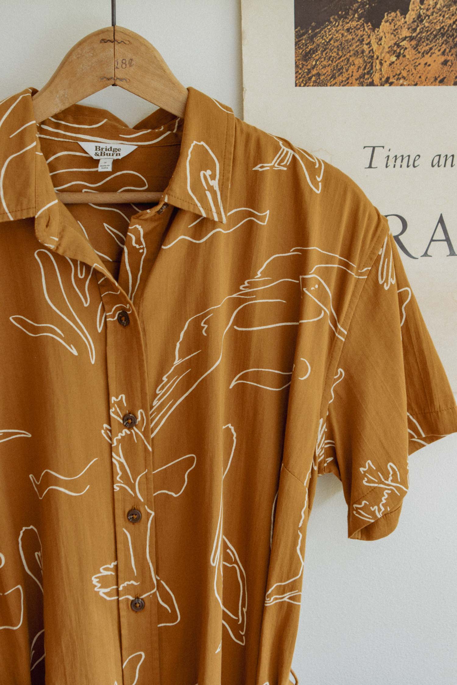 The Sylvie Dress / Shoreflight by Bridge & Burn, a brown short-sleeve button-up dress with white line art, hangs on a wooden hanger beside a partially visible poster on the wall.