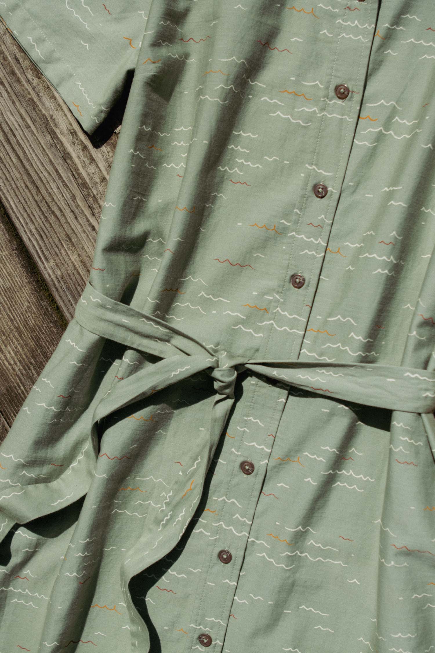 The Sylvie Dress by Bridge & Burn in Teal Tide Waves—an organic cotton shirtdress with a belt, short sleeves, and a subtle abstract line pattern—is displayed on a wooden surface in sunlight.