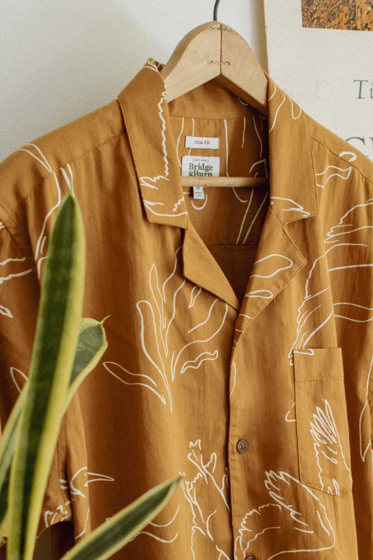 The Bridge & Burn Bodie Shirt in Shoreflight, a lightweight summer shirt featuring a white abstract leaf pattern on mustard yellow, hangs on a wooden hanger with a green plant in front, highlighting its retro camp collar and relaxed vibe.