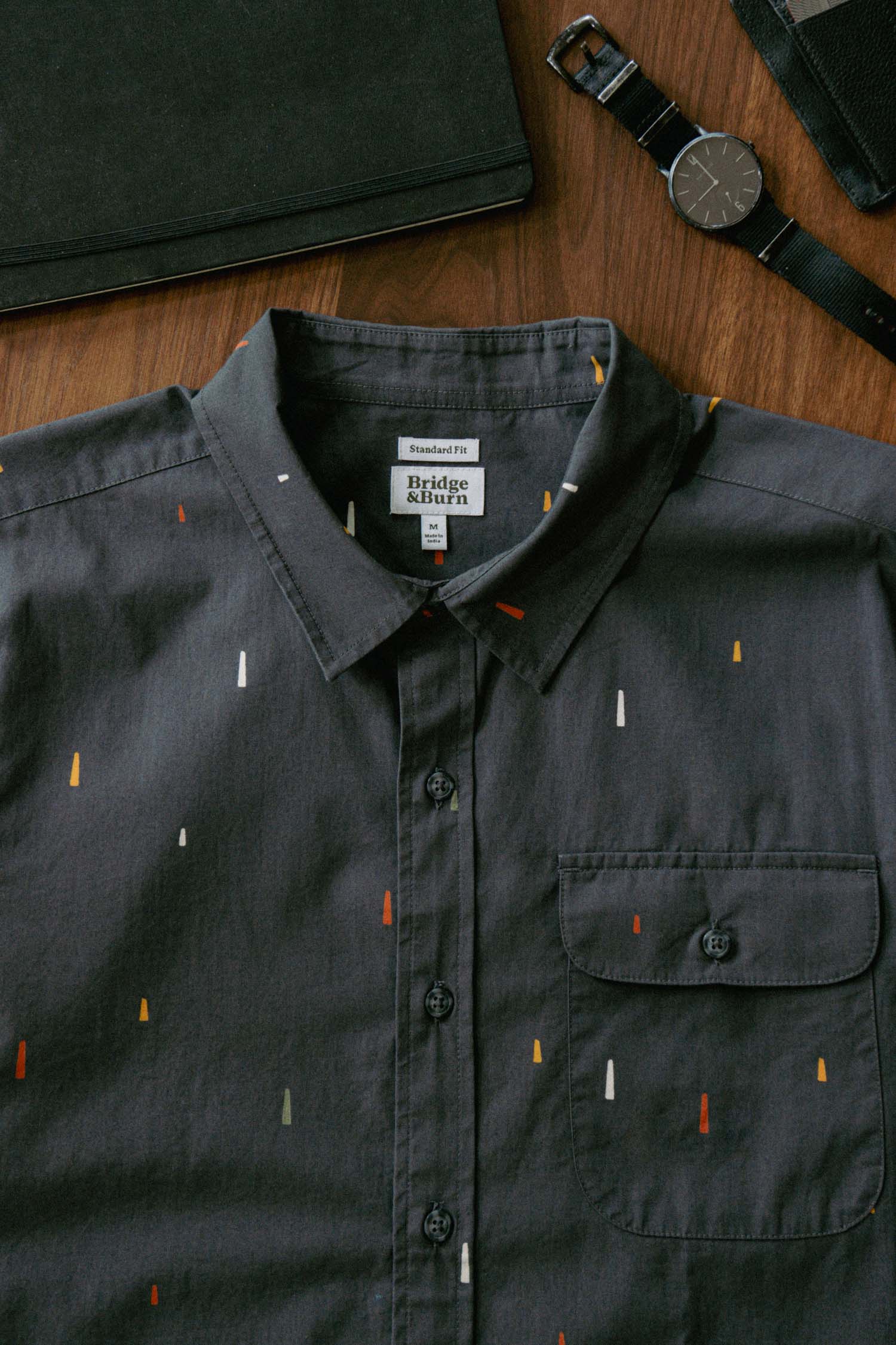 The Bridge & Burn Marten Shirt in Midnight Rain, a dark cotton casual shirt with a front pocket and multicolored dash patterns, is displayed on a wooden surface beside a black watch and folder.