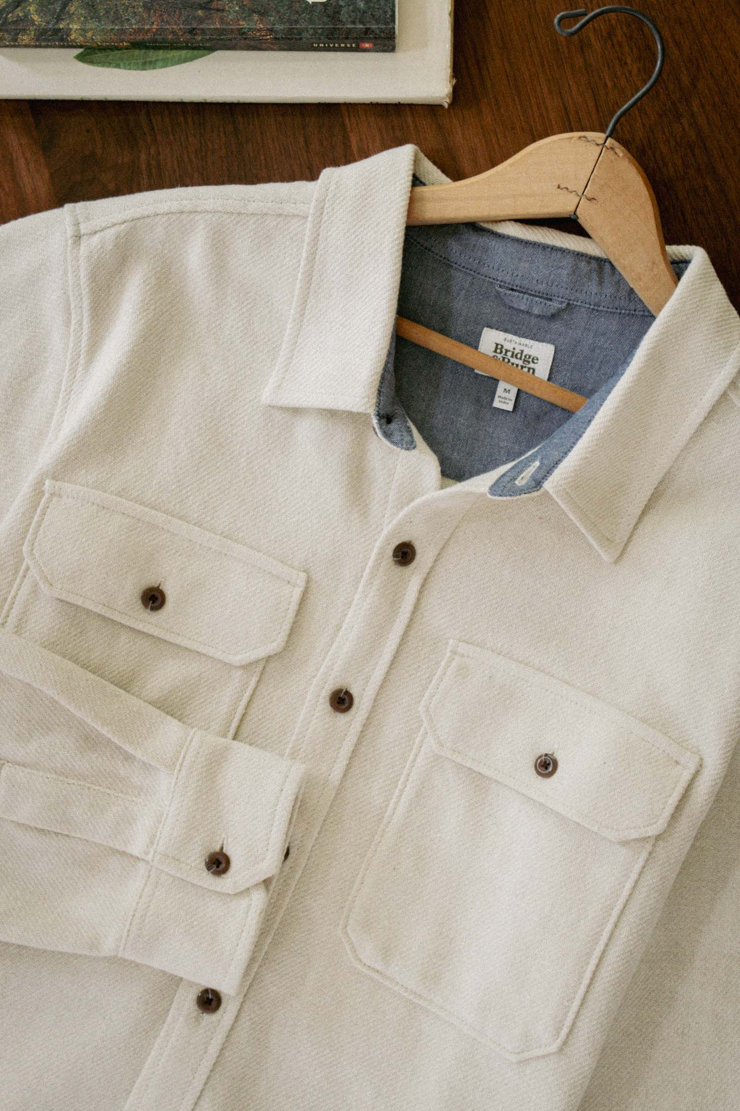 The Pointer Overshirt in ivory twill by Bridge & Burn features two chest pockets and is shown on a wooden hanger atop a wooden surface, reflecting the brand’s focus on sustainable clothing.