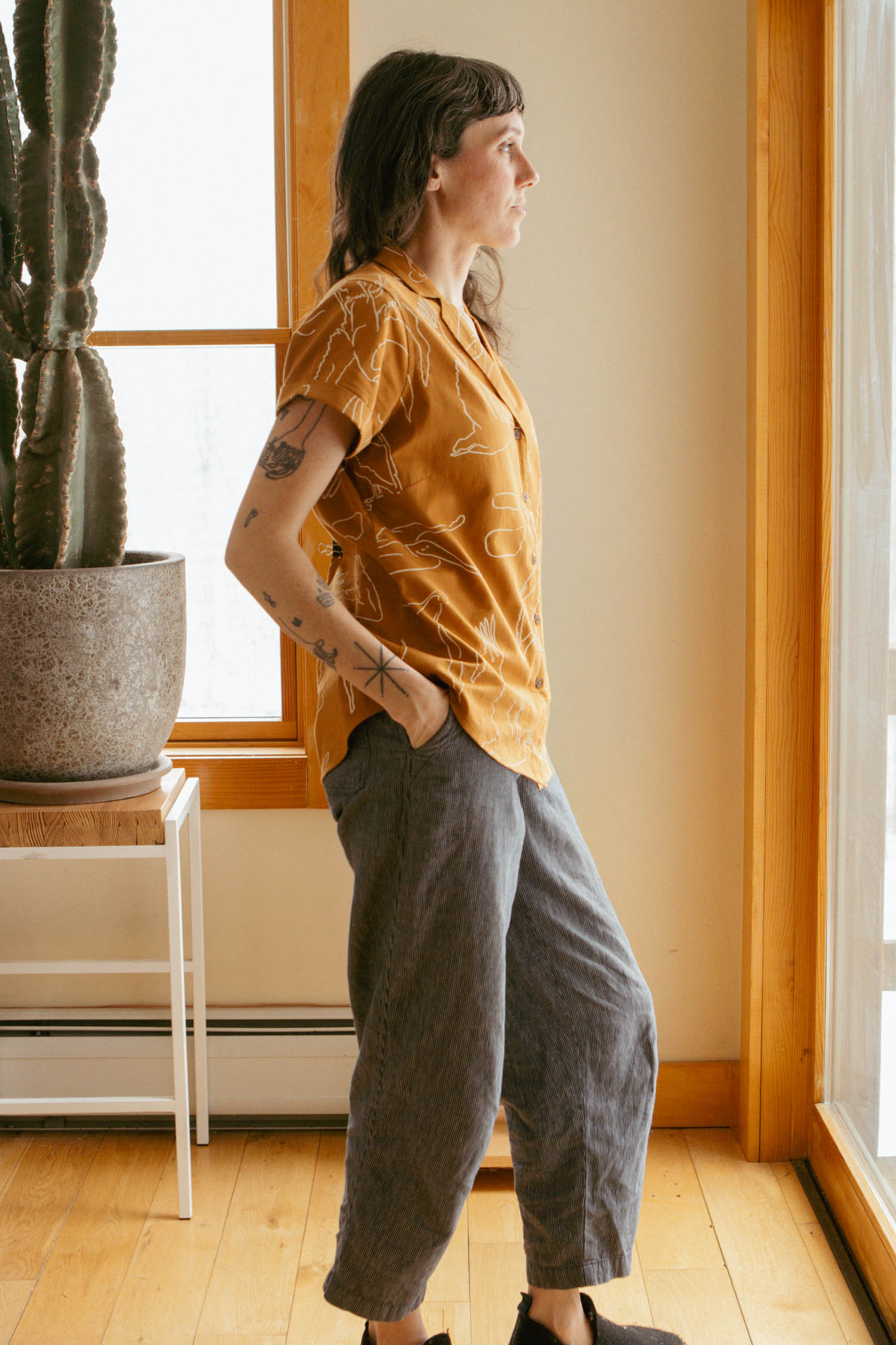 A person with shoulder-length hair stands by a window, hands in pockets, wearing the Bridge & Burn Innes Shirt in Shoreflight and gray pants, next to a potted cactus indoors.