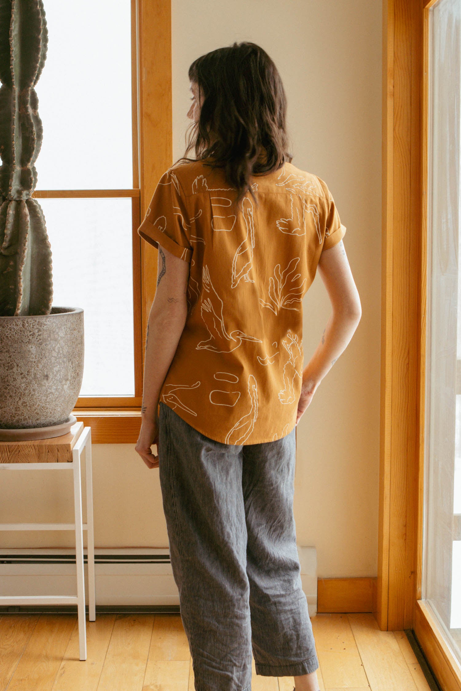 A person with shoulder-length dark hair stands indoors facing away, wearing Bridge & Burn’s relaxed fit Innes Shirt in Shoreflight and loose gray pants. A large potted cactus sits nearby near a window.