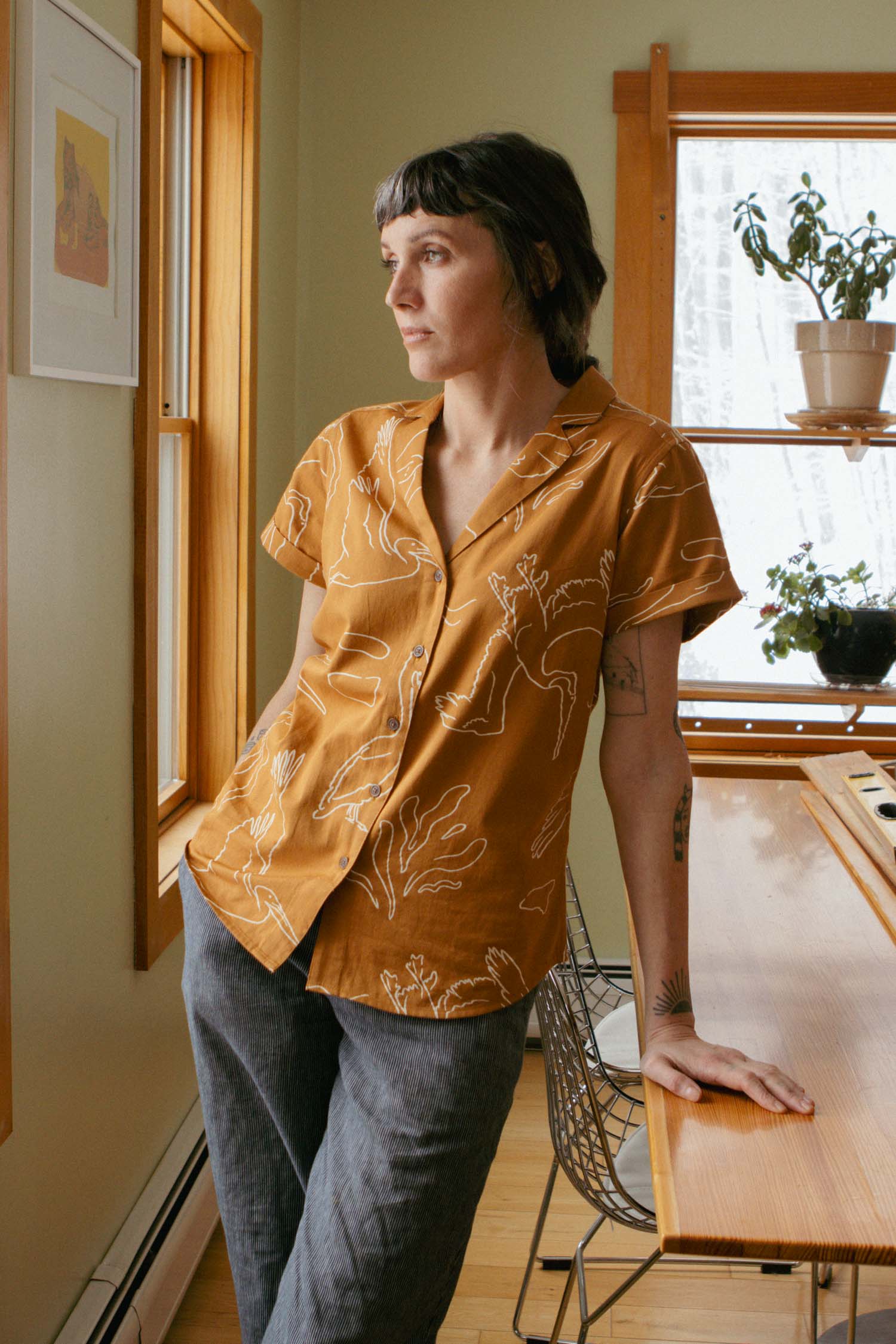 A person with short brown hair wears the Bridge & Burn Innes Shirt in Shoreflight and gray pants, leaning against a wooden table indoors and gazing thoughtfully out a window.
