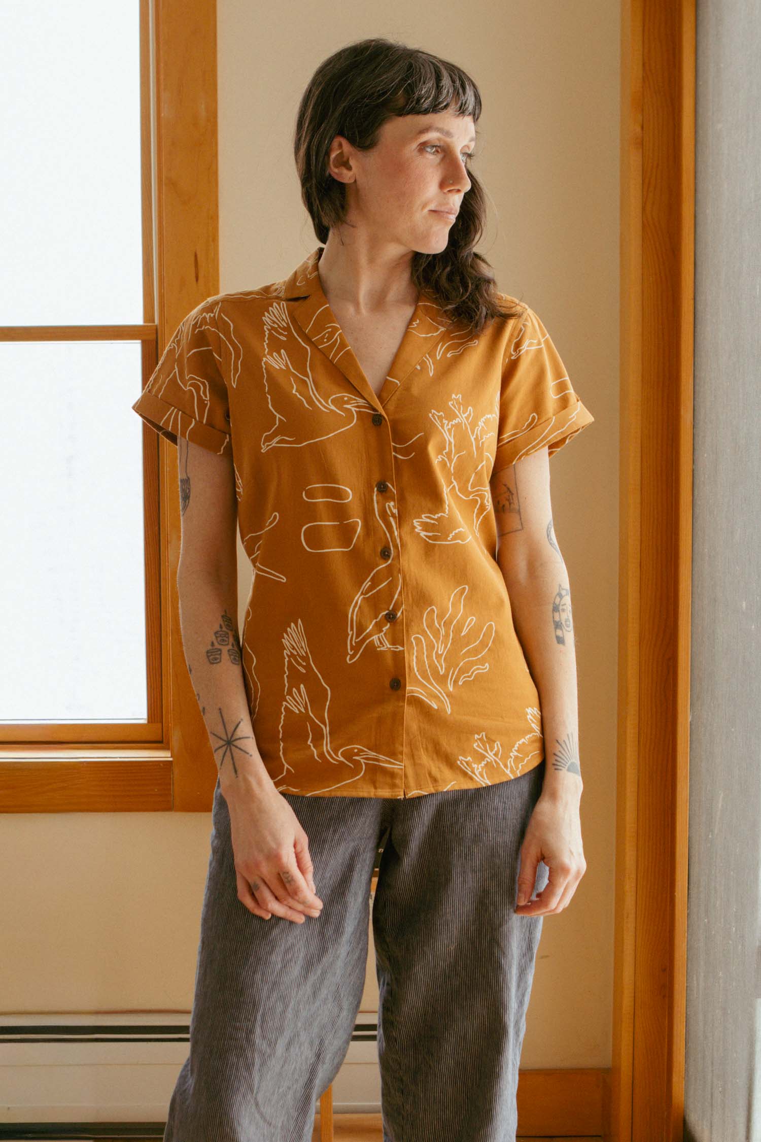 A person with long brown hair and tattoos stands indoors by a window, wearing the Bridge & Burn Innes Shirt in Shoreflight—a mustard yellow organic cotton top with white animal patterns—styled casually with loose gray pants.