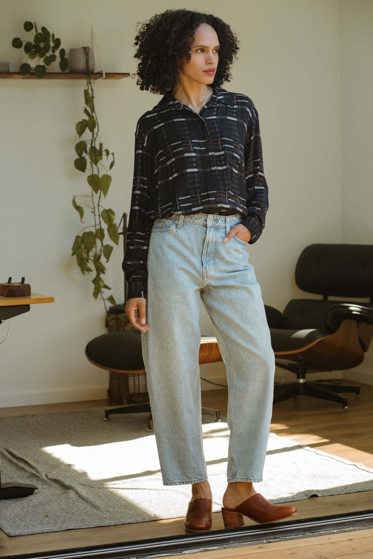 A person with curly hair wears a Bridge & Burn Haley Shirt in Nightfall Stripe, relaxed fit, paired with light blue high-waisted jeans and brown shoes, standing in a sunlit room with modern furniture and a wall-mounted plant.