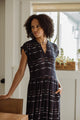 A woman with curly hair stands indoors by a window, wearing the Bridge & Burn Ira Dress in Nightfall Stripe. She looks to the side with one hand on a wooden chair as sunlight streams through behind her.