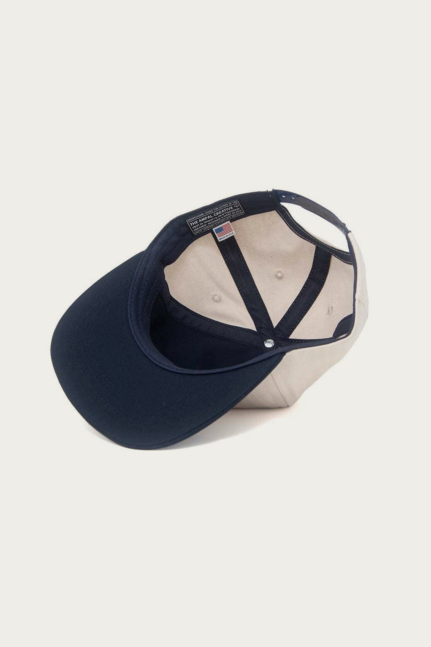The Ampal Creative - Don't Think Twice Snapback in navy is shown upside down, displaying its inner lining, seam details, an American flag tag, and a distinctive Ampal patch inside.