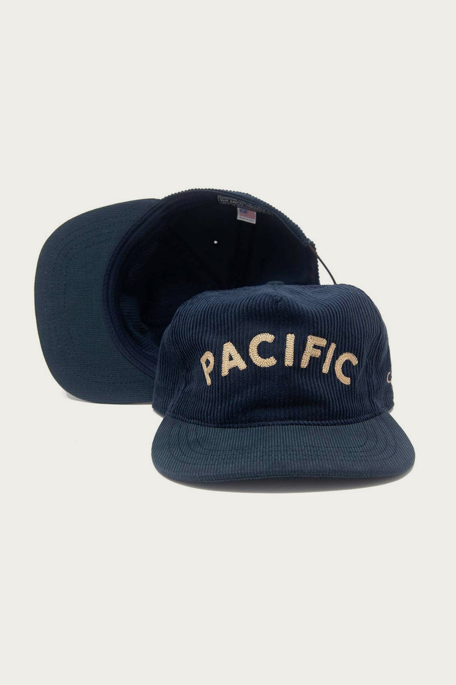 Two navy blue The Ampal Creative - Pacific III Strapback hats are shown: one upright with “PACIFIC” in cream vintage-style embroidery, the other lying flat behind—a must-have for classic style fans.