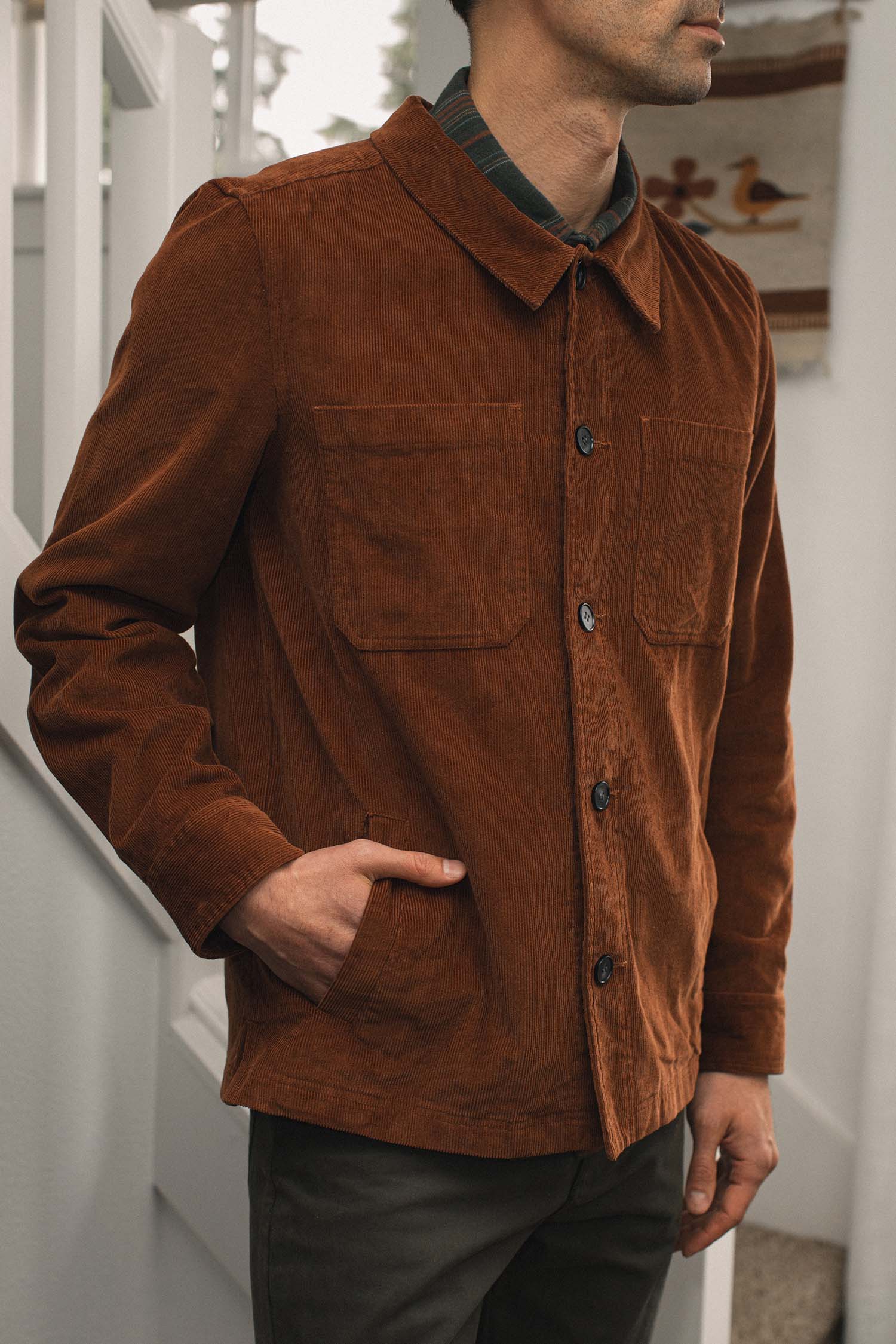 A man wears the Bridge & Burn Banks Shirt Jacket in Saddle Brown, featuring two chest pockets, standing indoors with one hand in his pocket. His head is partially out of frame, with white stairs and a patterned wall hanging behind him.