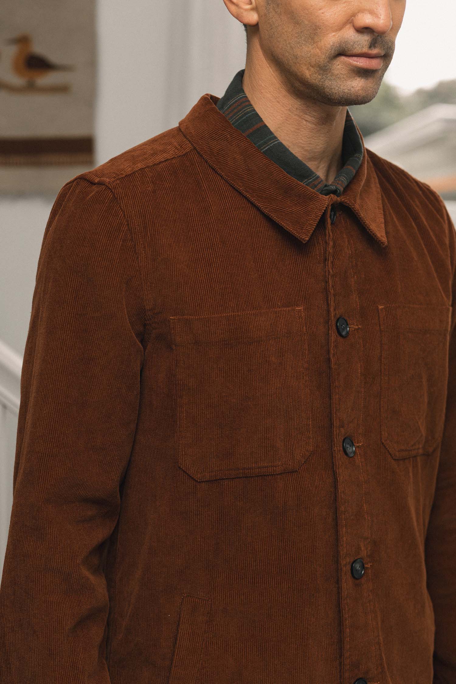 A man wearing the Bridge & Burn Banks Shirt Jacket in Saddle Brown, layered over a collared shirt, stands indoors with his face partially out of frame. The jacket features large front pockets and a corduroy texture.