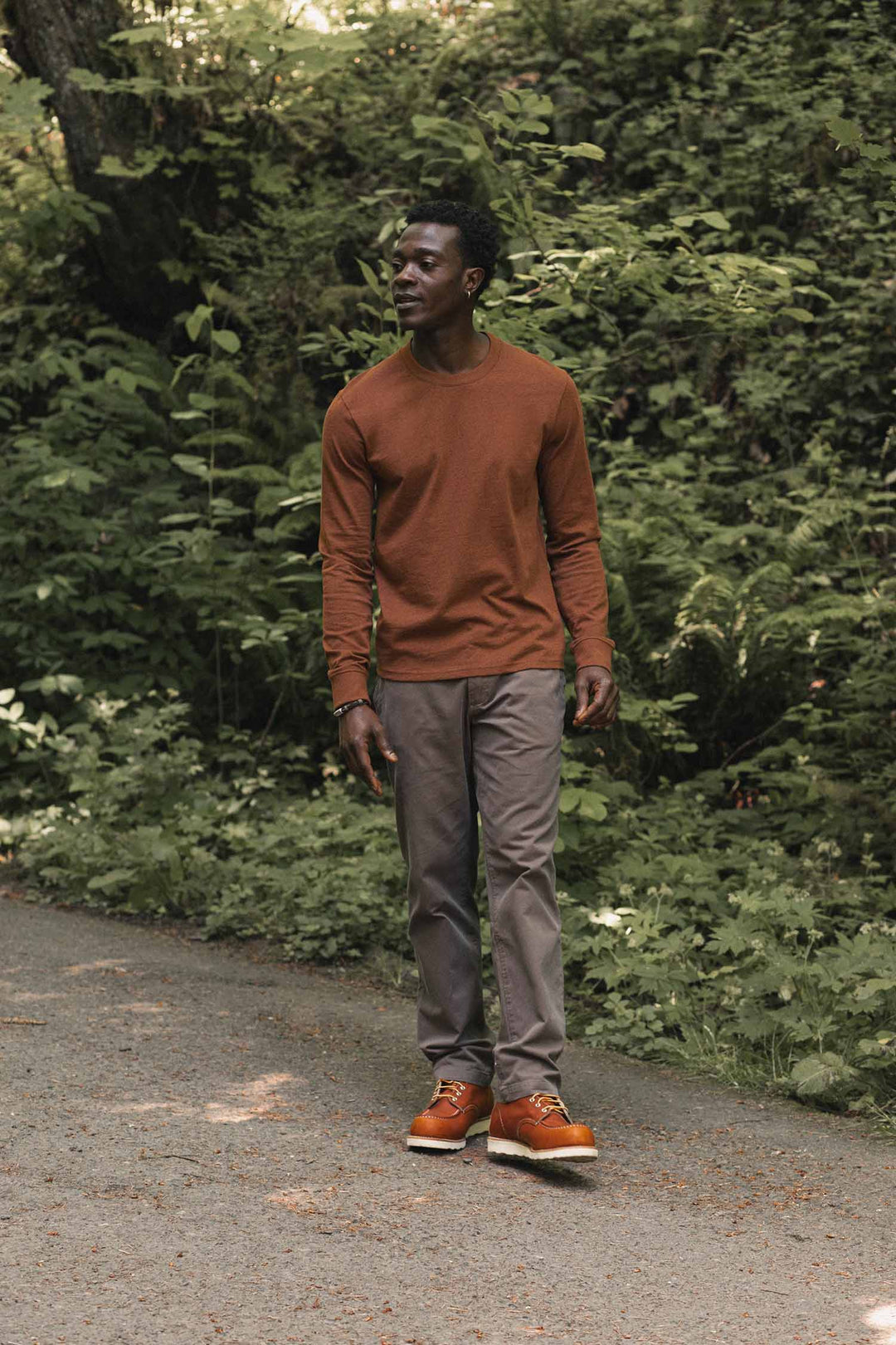A man wearing the Bridge & Burn Barney Long Sleeve Tee in Bronze, along with gray pants and orange shoes, walks on a paved path surrounded by lush green foliage.