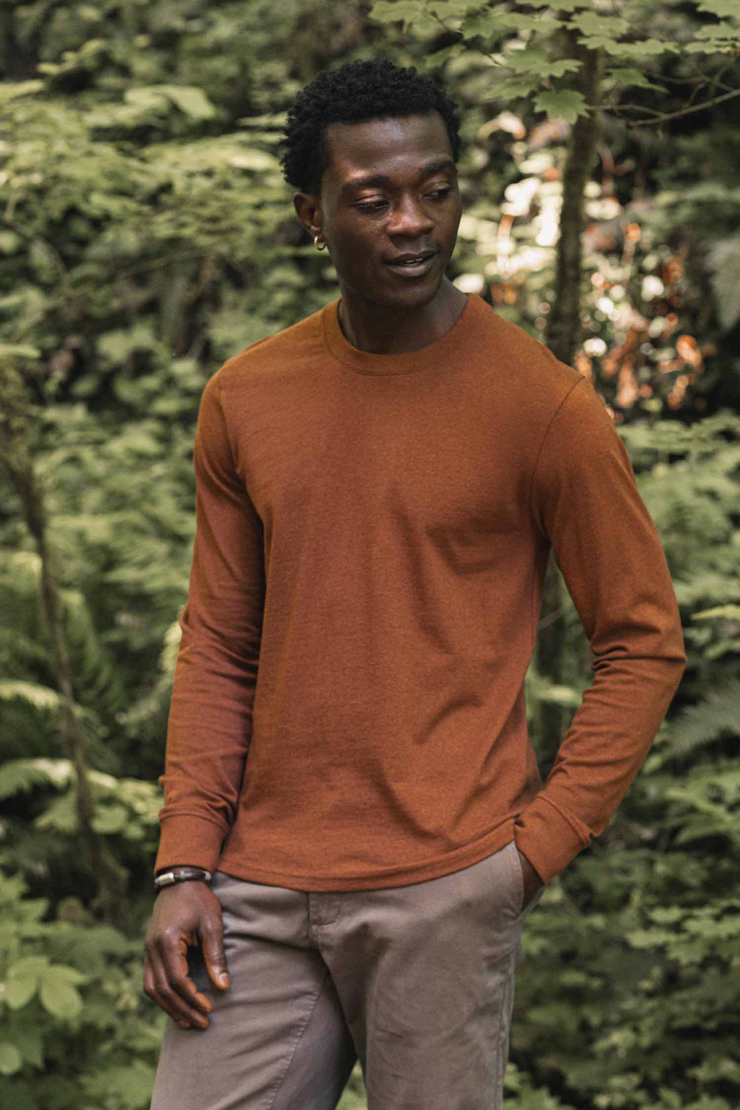 A man wearing the Bridge & Burn Barney Long Sleeve Tee in Bronze and gray pants stands outdoors in a lush, green forest, looking slightly to the side.
