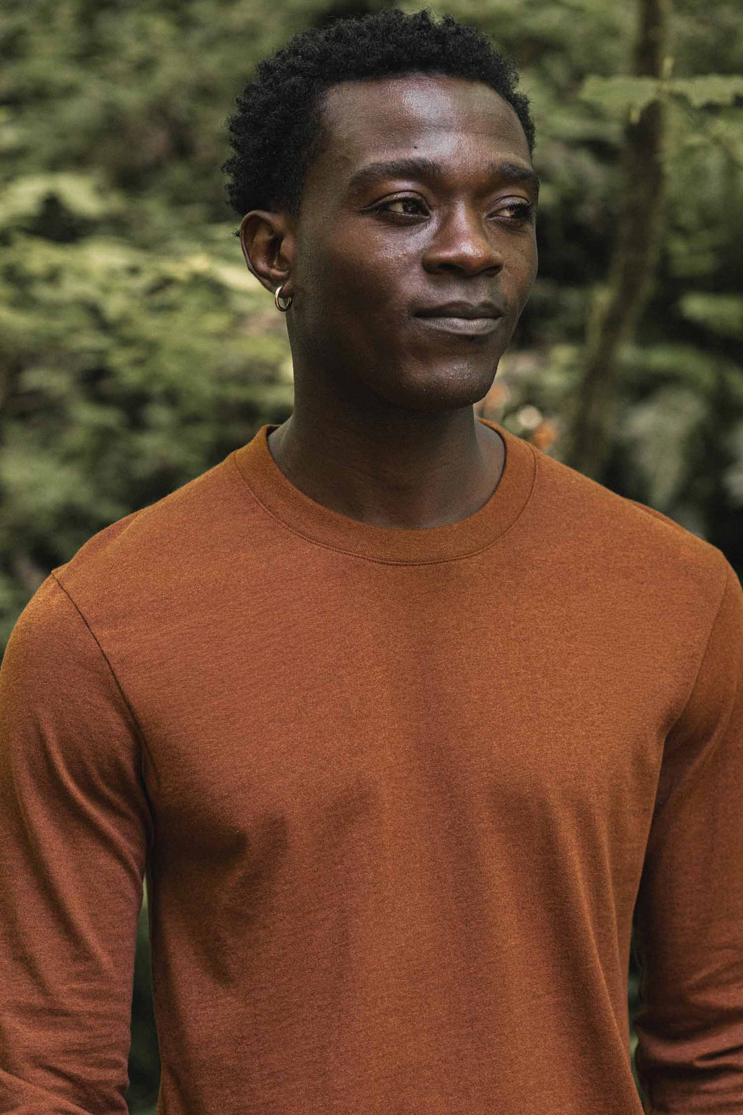 A person wearing the Bridge & Burn Barney Long Sleeve Tee in Bronze, featuring a relaxed fit and crew neck design, stands outdoors in front of green foliage, looking slightly to the side with a neutral expression.
