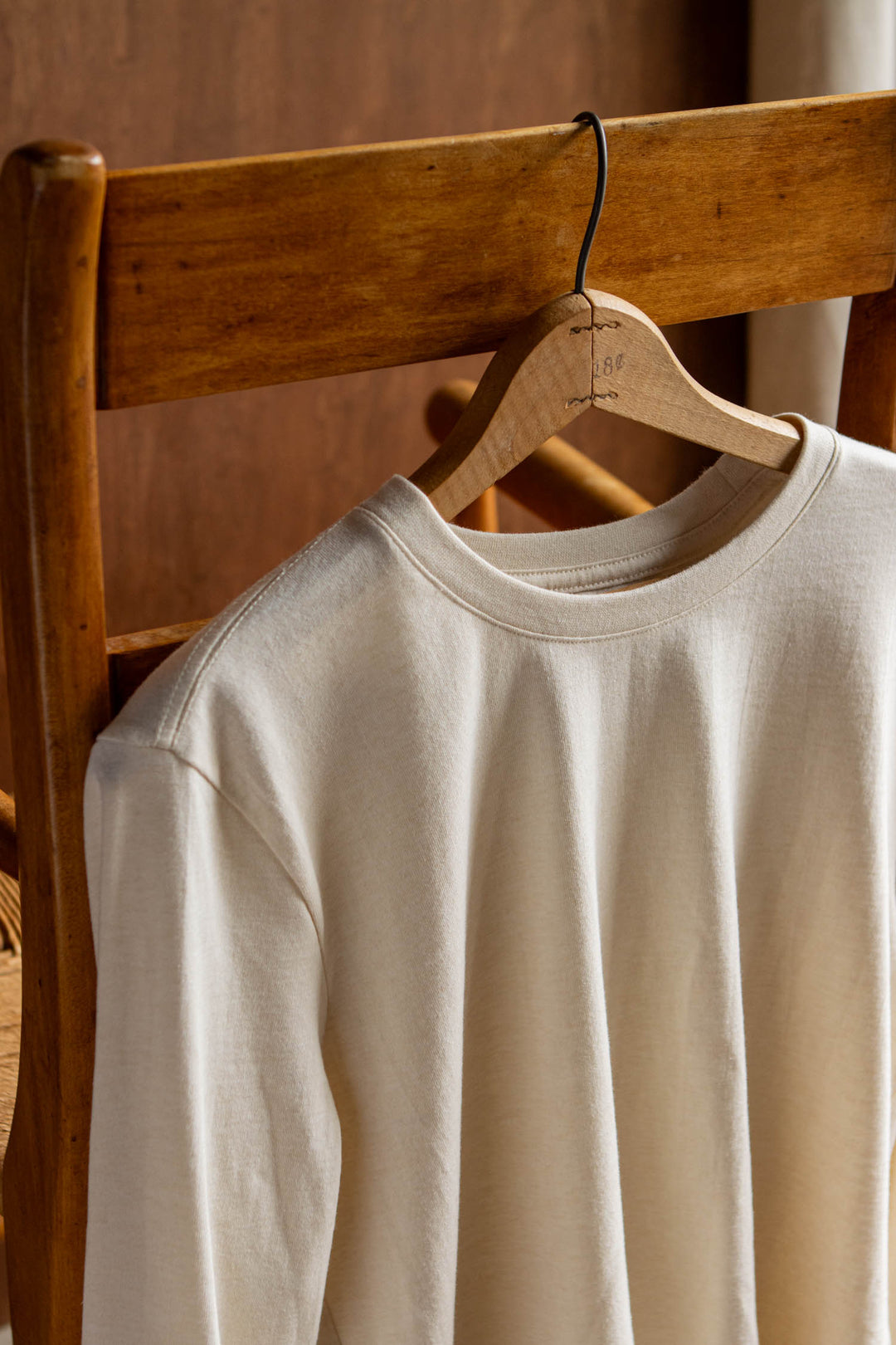 The Barney Long Sleeve Tee in Ivory by Bridge & Burn, featuring a relaxed fit and crew neck design, is draped over the back of a wooden chair on a wooden hanger, with soft natural light highlighting the recycled cotton fabric’s texture.