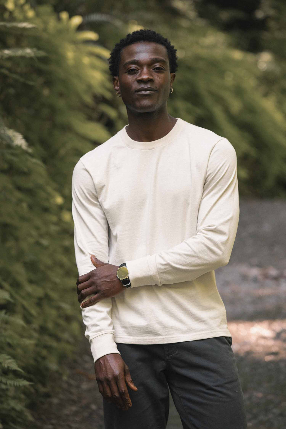 A man stands outdoors on a path surrounded by greenery, arms gently crossed and wearing the Bridge & Burn Barney Long Sleeve Tee in Ivory—a relaxed fit crew neck shirt crafted from recycled cotton—paired with dark pants and a relaxed expression.