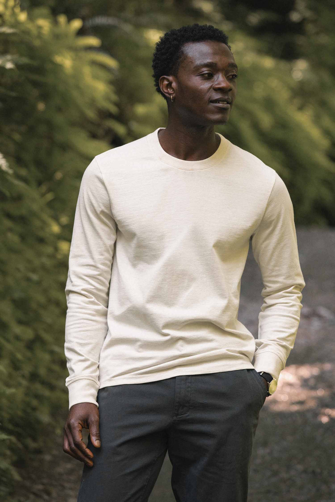 A man stands outdoors on a gravel path, wearing the Barney Long Sleeve Tee in Ivory by Bridge & Burn paired with dark pants, surrounded by greenery. With one hand in his pocket and looking to the side, he exudes a relaxed confidence in this effortlessly stylish, relaxed fit look.