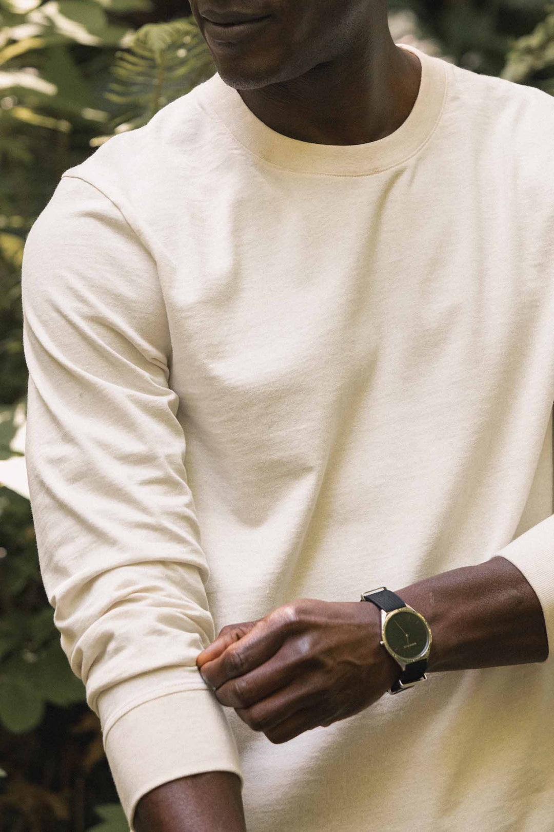 A person wearing the Bridge & Burn Barney Long Sleeve Tee in Ivory, a relaxed fit crew neck shirt crafted from recycled cotton, stands outdoors with their sleeve rolled up and a black wristwatch on their wrist. Green foliage is visible in the background.