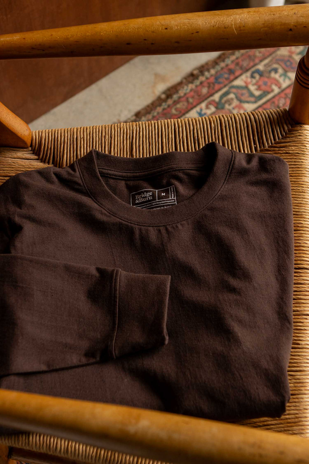 A folded Barney Long Sleeve Tee in Truffle by Bridge & Burn, featuring a relaxed fit and crew neck with a visible label, rests on a woven wooden chair; part of a patterned rug and the floor can be seen in the background.