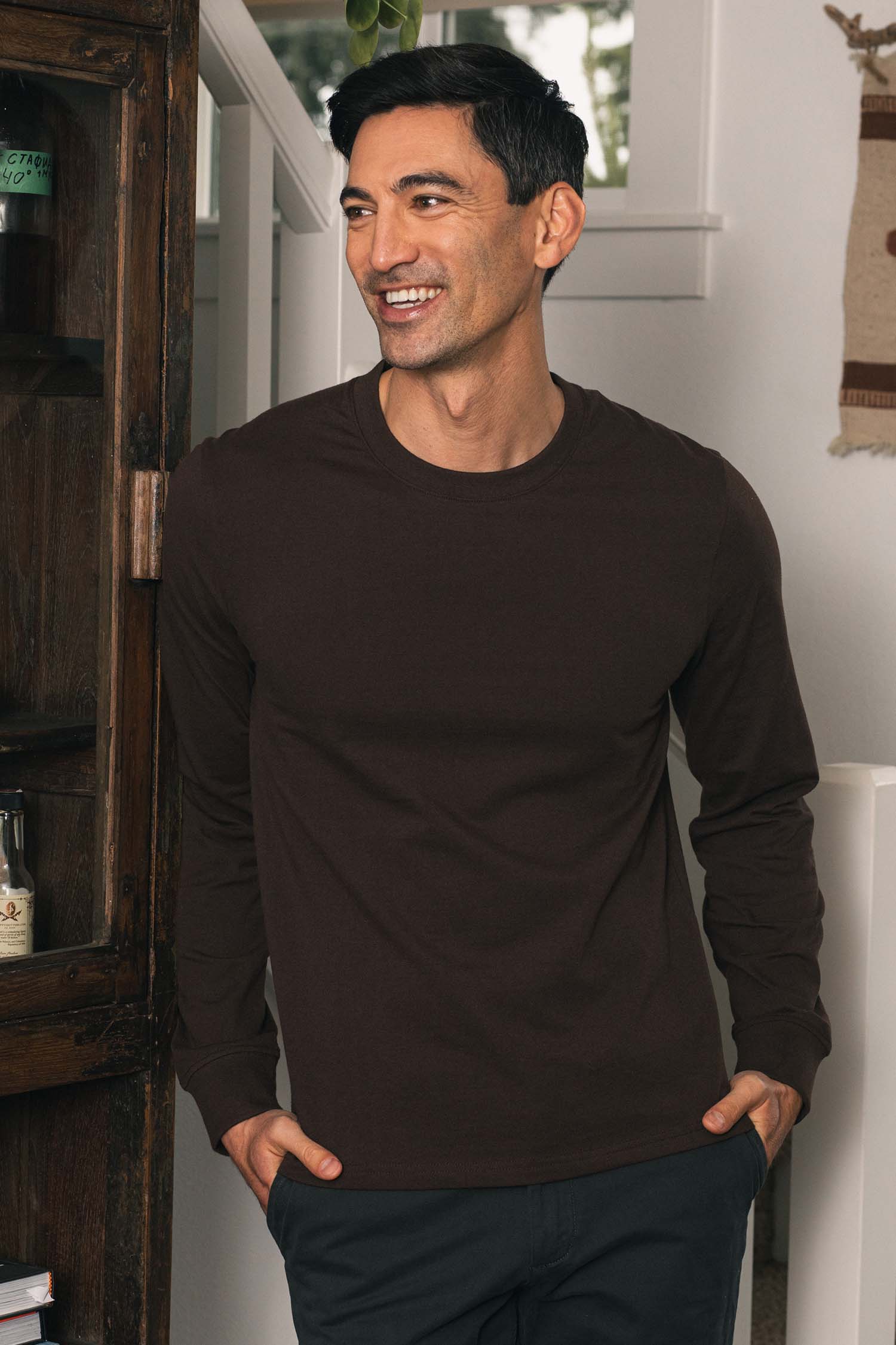 A man stands indoors, smiling and looking to the side with his hands in his pockets. He is wearing the Bridge & Burn Barney Long Sleeve Tee in Truffle— a dark brown, crew neck shirt made from recycled cotton—paired with relaxed fit dark pants. In the background, there is a wooden cabinet and a wall hanging.