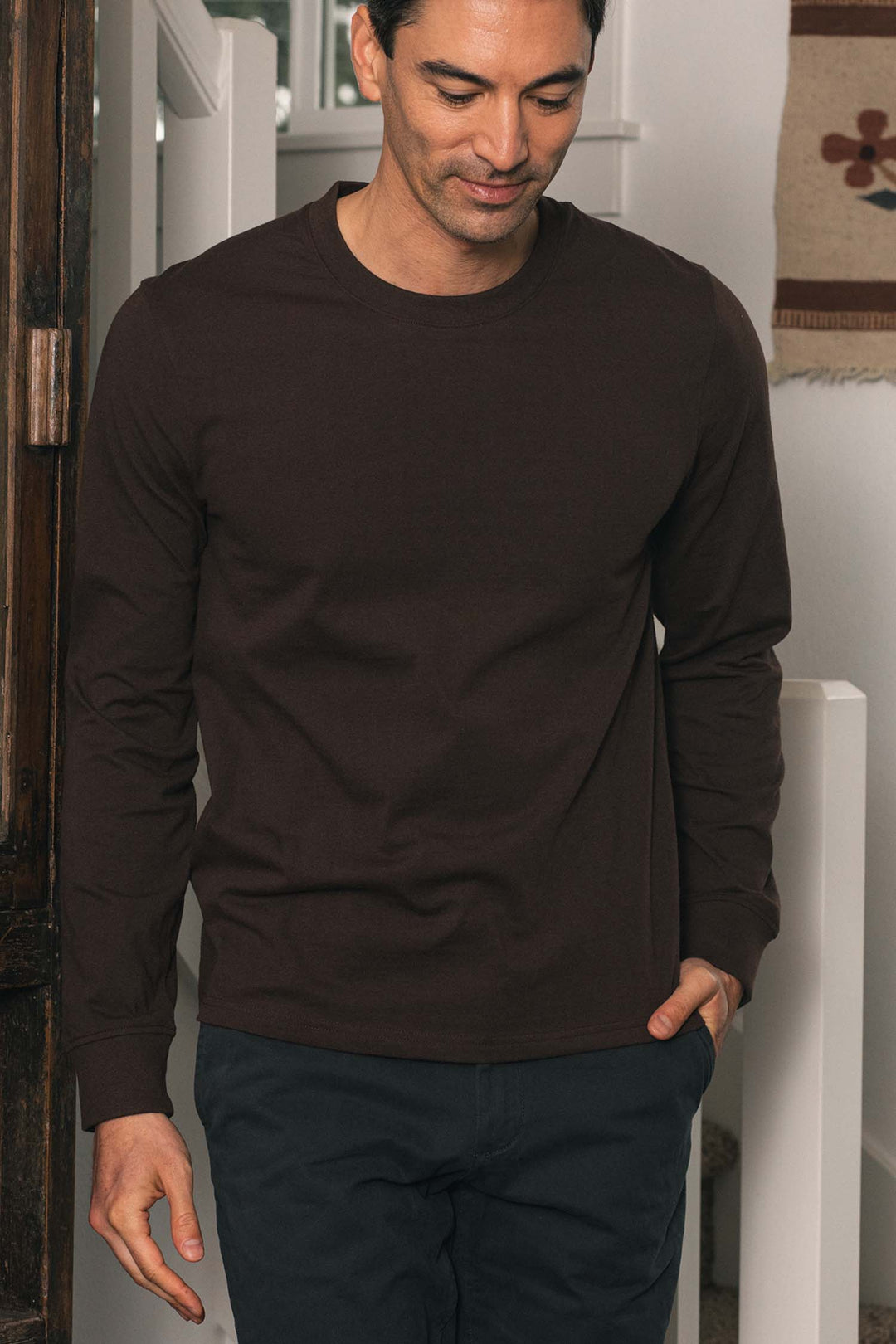 A man wearing the Bridge & Burn Barney Long Sleeve Tee in Truffle—a dark brown, relaxed fit crew neck shirt crafted from recycled cotton—stands indoors beside a wooden door and white stairs, looking down with a slight smile and one hand in his pocket.