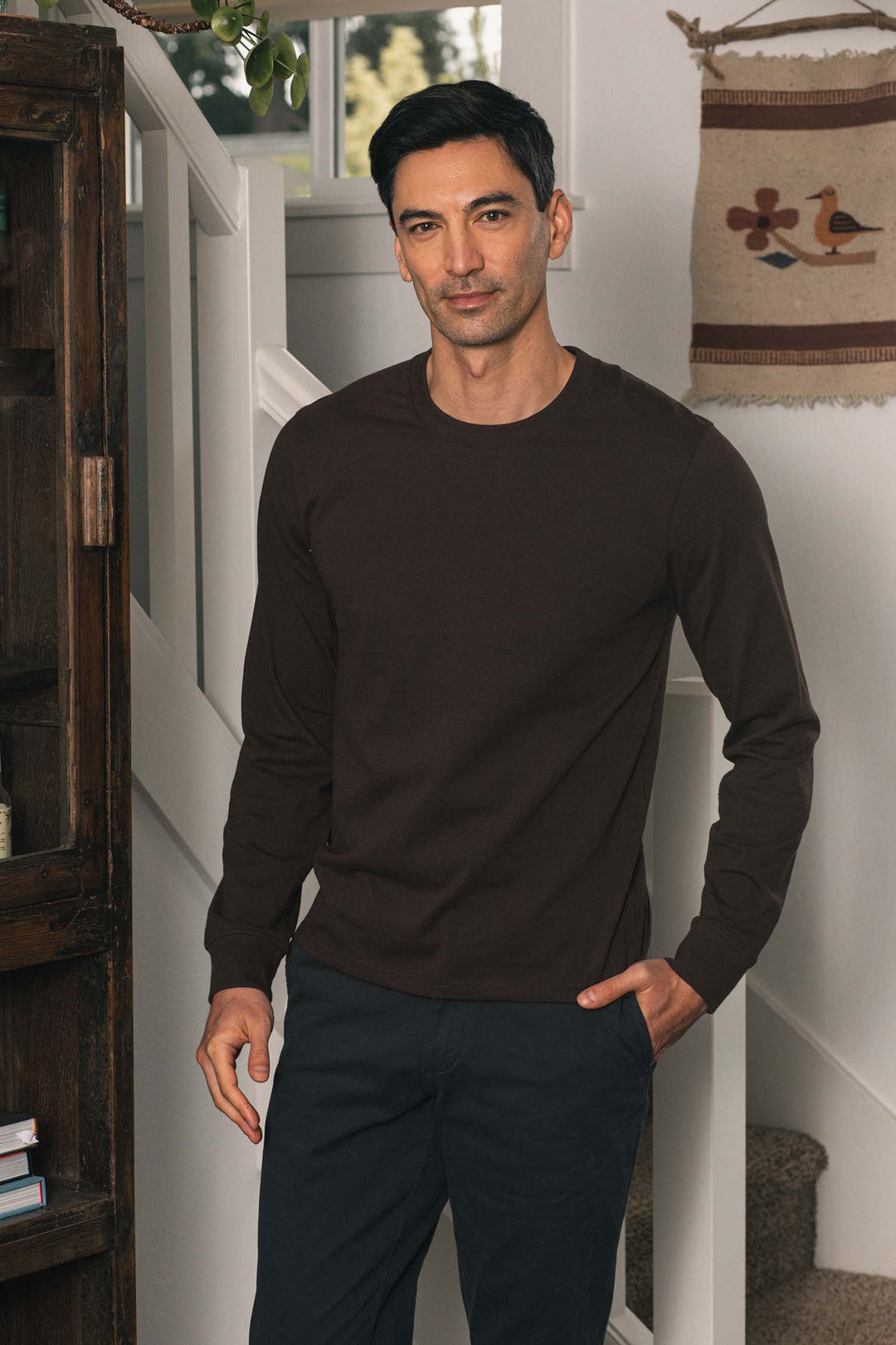 A man with short dark hair wears the Bridge & Burn Barney Long Sleeve Tee in Truffle and dark pants, standing indoors near a white staircase and a wooden cabinet, with a woven wall hanging in the background.