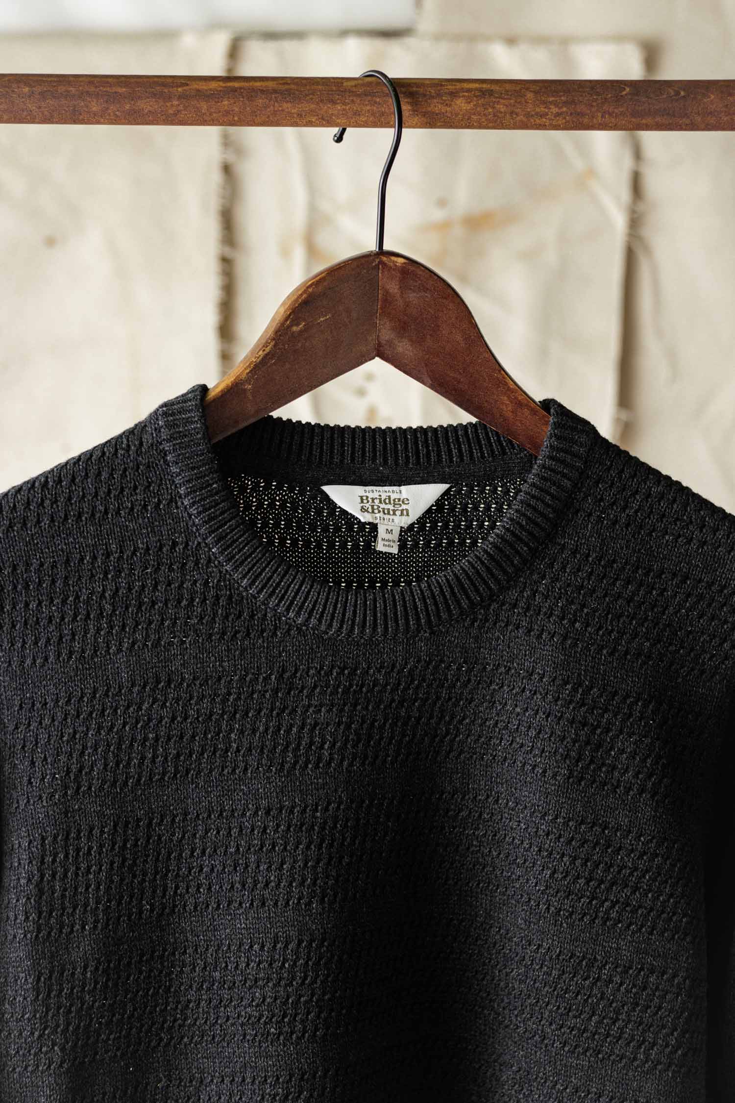 The Bridge & Burn Calder Sweater in Midnight, featuring a black textured sustainable design, hangs on a wooden hanger against a neutral, slightly wrinkled background.