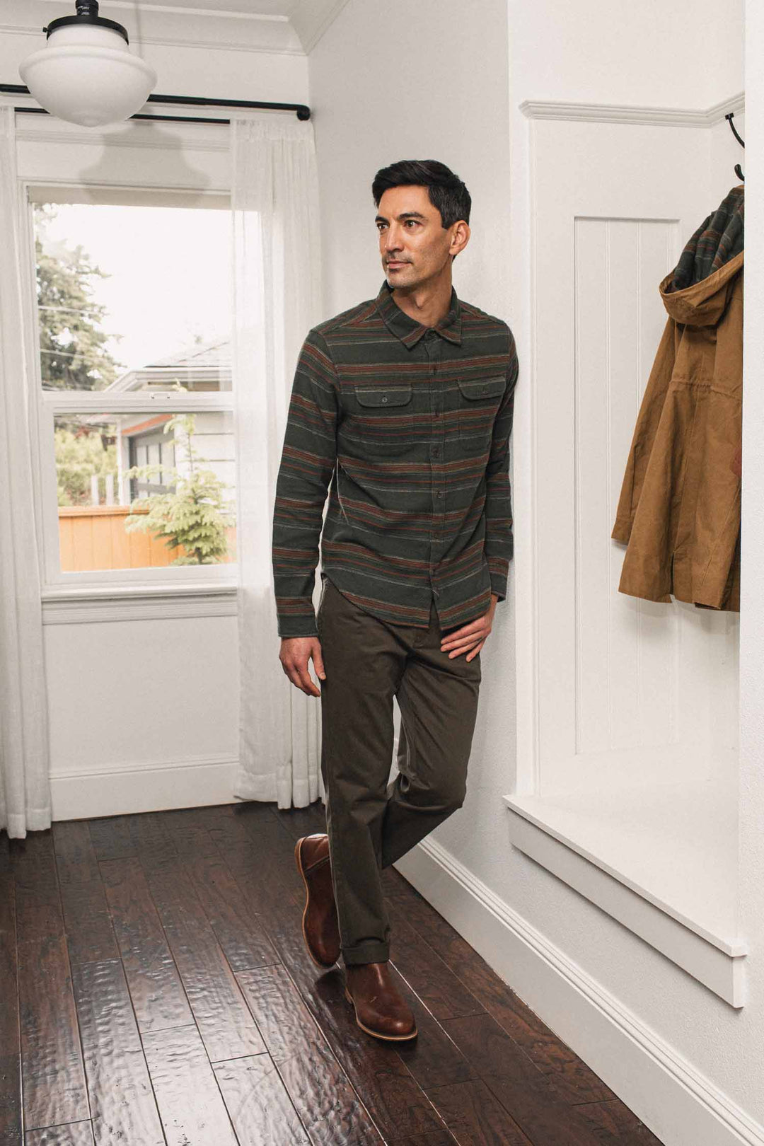 A man wears a Bridge & Burn Cole Slim Shirt / Mallard Stripe, a long-sleeved, slim-fit button-down with horizontal multi-colored stripes, as he leans against a white wall in dark trousers.