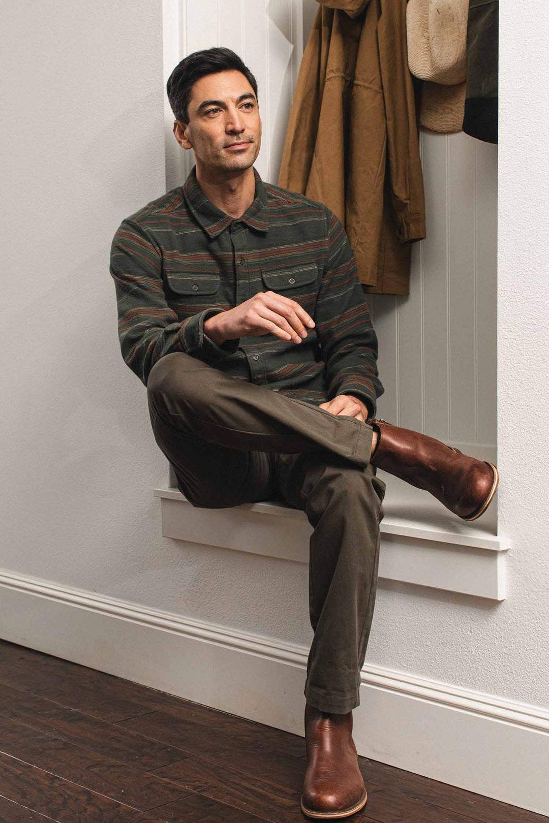A man wearing a Bridge & Burn Cole Slim Shirt / Mallard Stripe with horizontal red and tan stripes and olive trousers sits on a window ledge, wearing brown leather boots.
