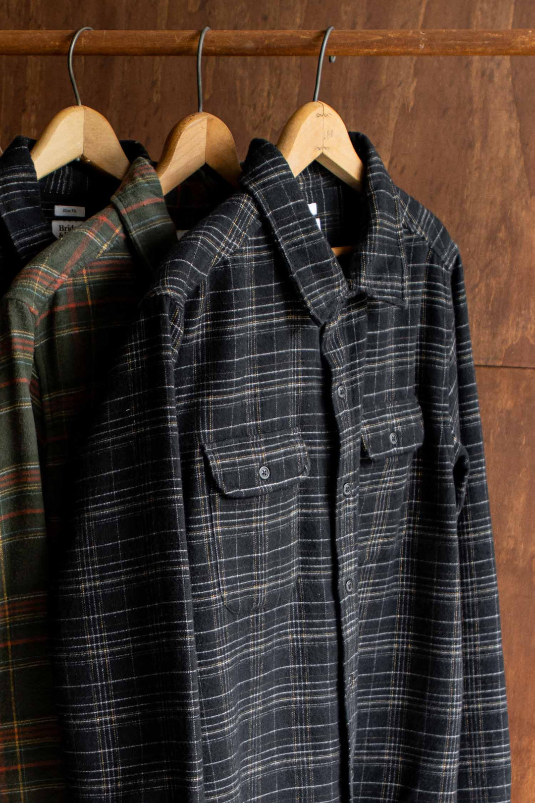 Three Cole Slim Shirt / Obsidian Grid long-sleeved, brushed cotton button-down shirts with plaid patterns hanging on wooden hangers.