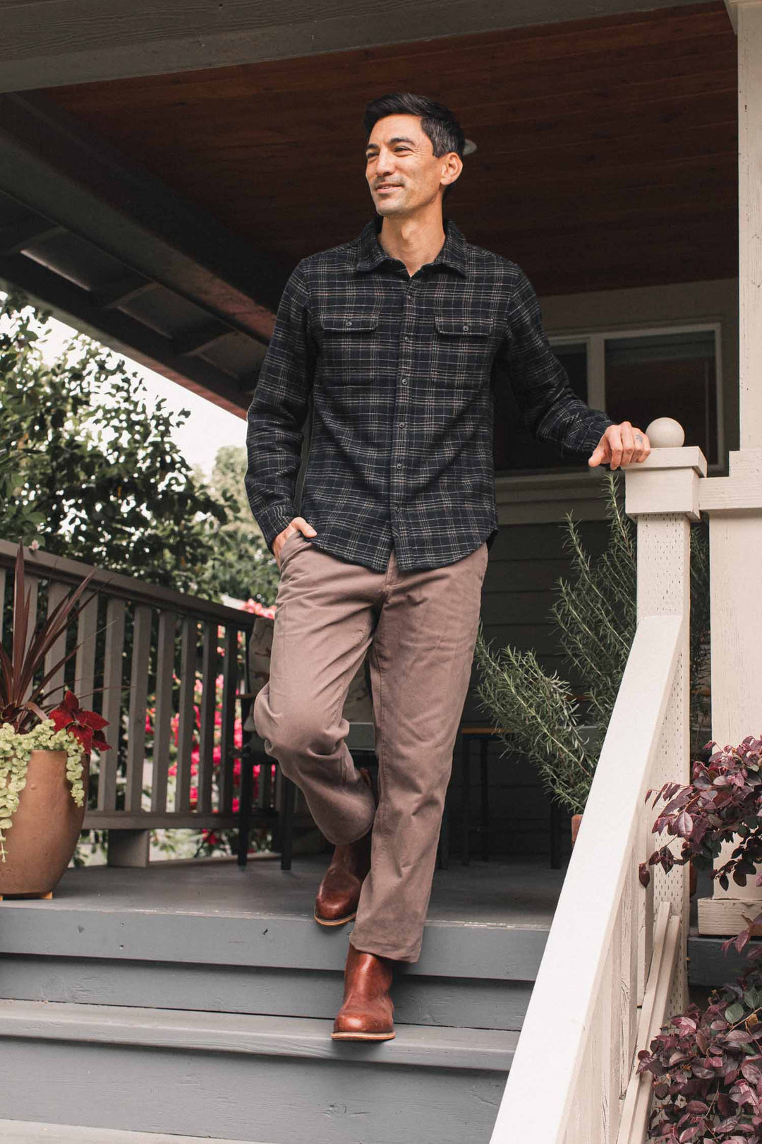 A man standing on a porch wearing a Bridge & Burn Cole Slim Shirt / Obsidian Grid, brown trousers, and brown leather boots.