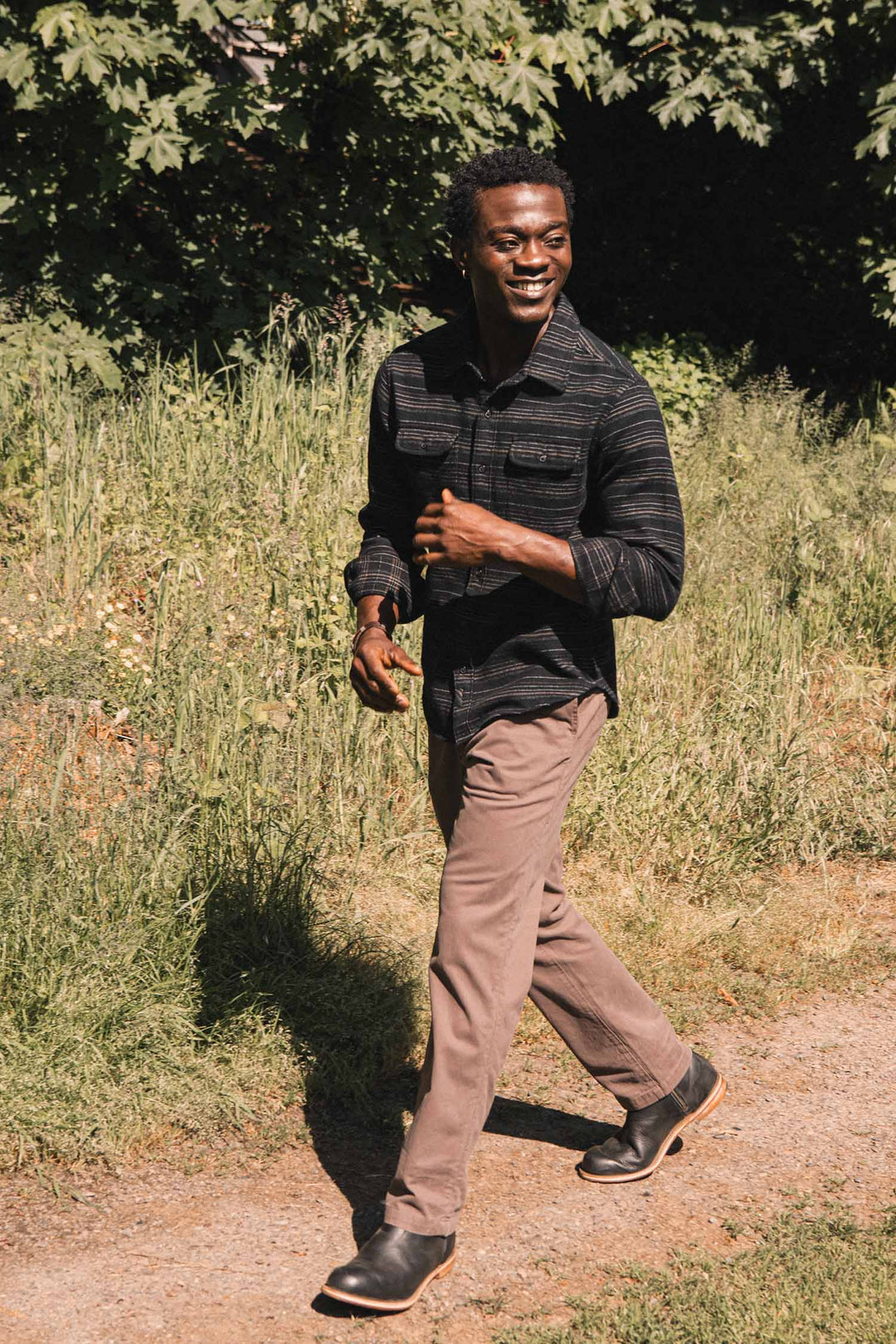 A man smiles while walking outside wearing a Bridge & Burn Cole Slim Shirt / Raven Stripe, a long-sleeved black button-down with thin horizontal stripes, paired with brown pants.