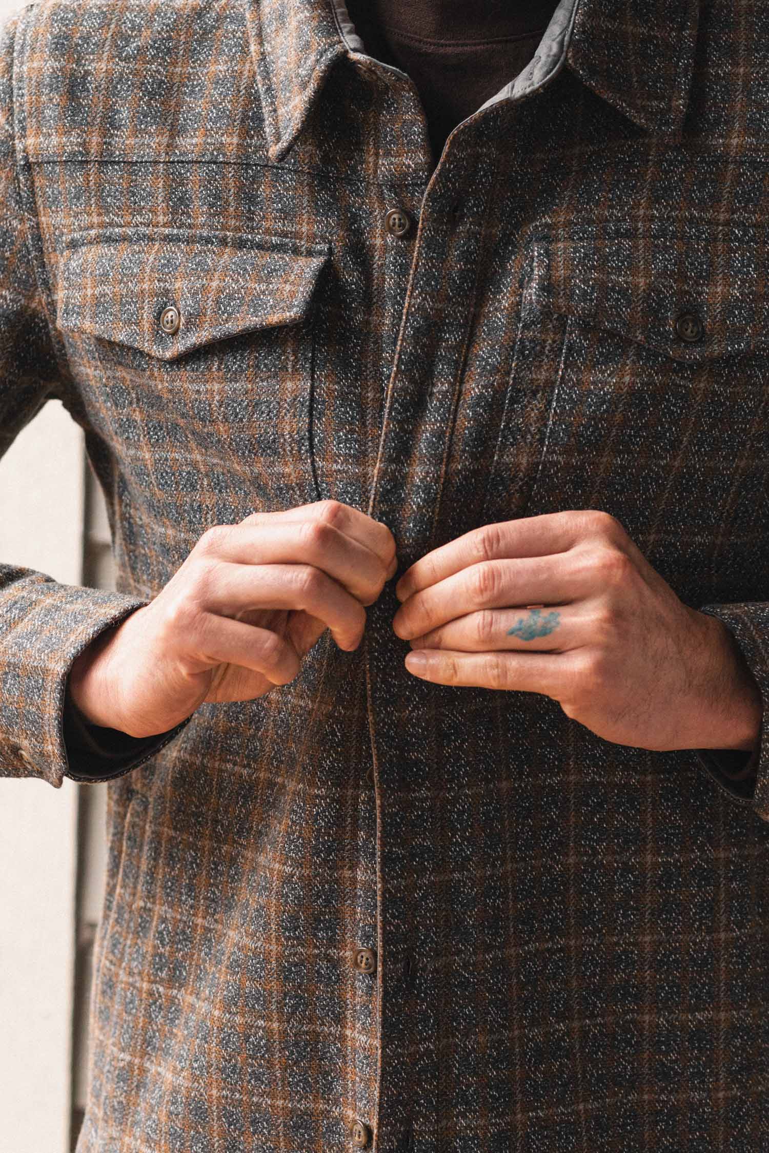 Someone buttons up the Bridge & Burn Fielding Shirt Jacket in Hearth Plaid, showing their hands—one with a small blue tattoo. The photo centers on the workwear-inspired flannel jacket and hands.