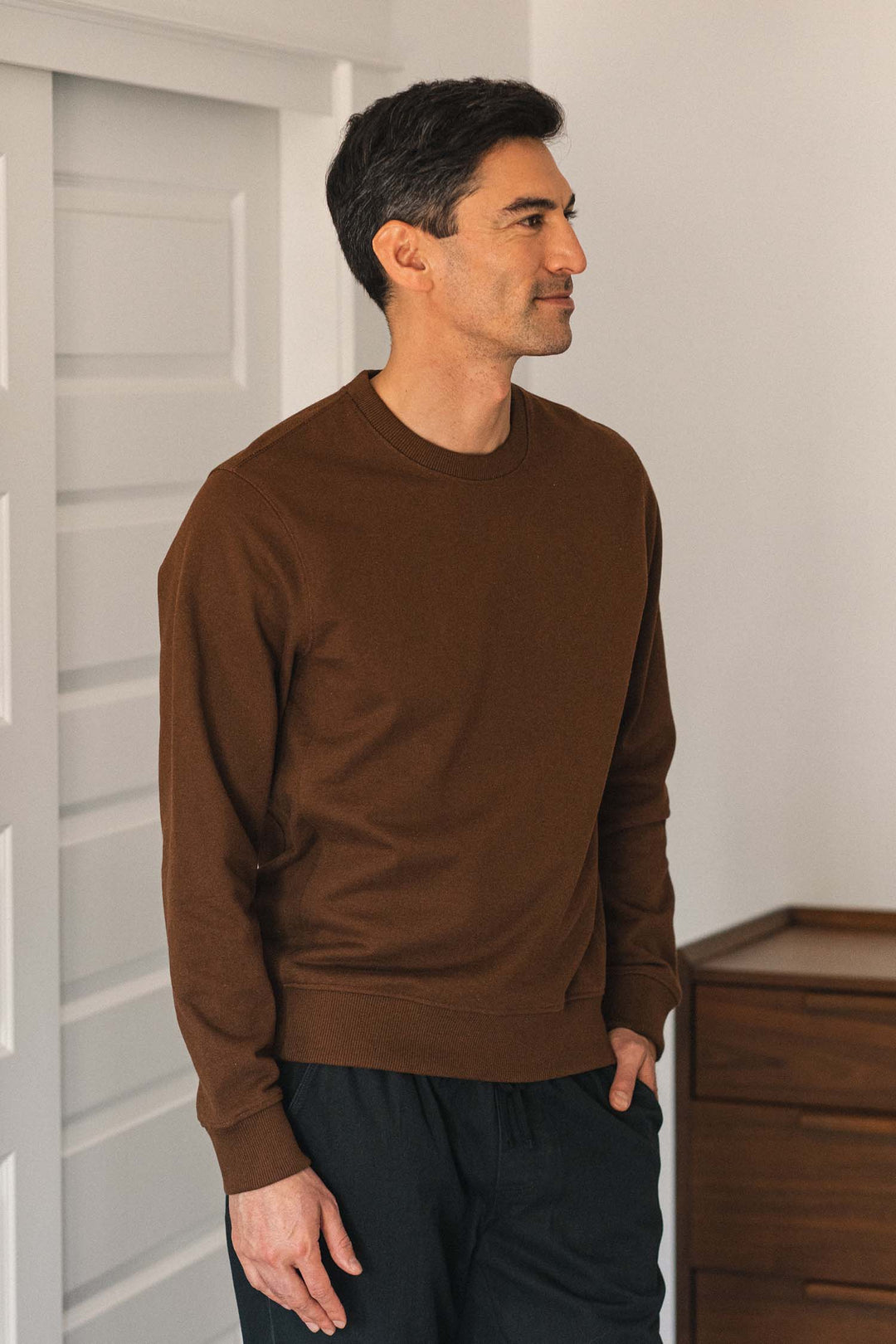 A man with short dark hair stands indoors with one hand in his pocket, looking to the side, wearing the Bridge & Burn Hayden Crew Sweatshirt in Cumin and black pants.