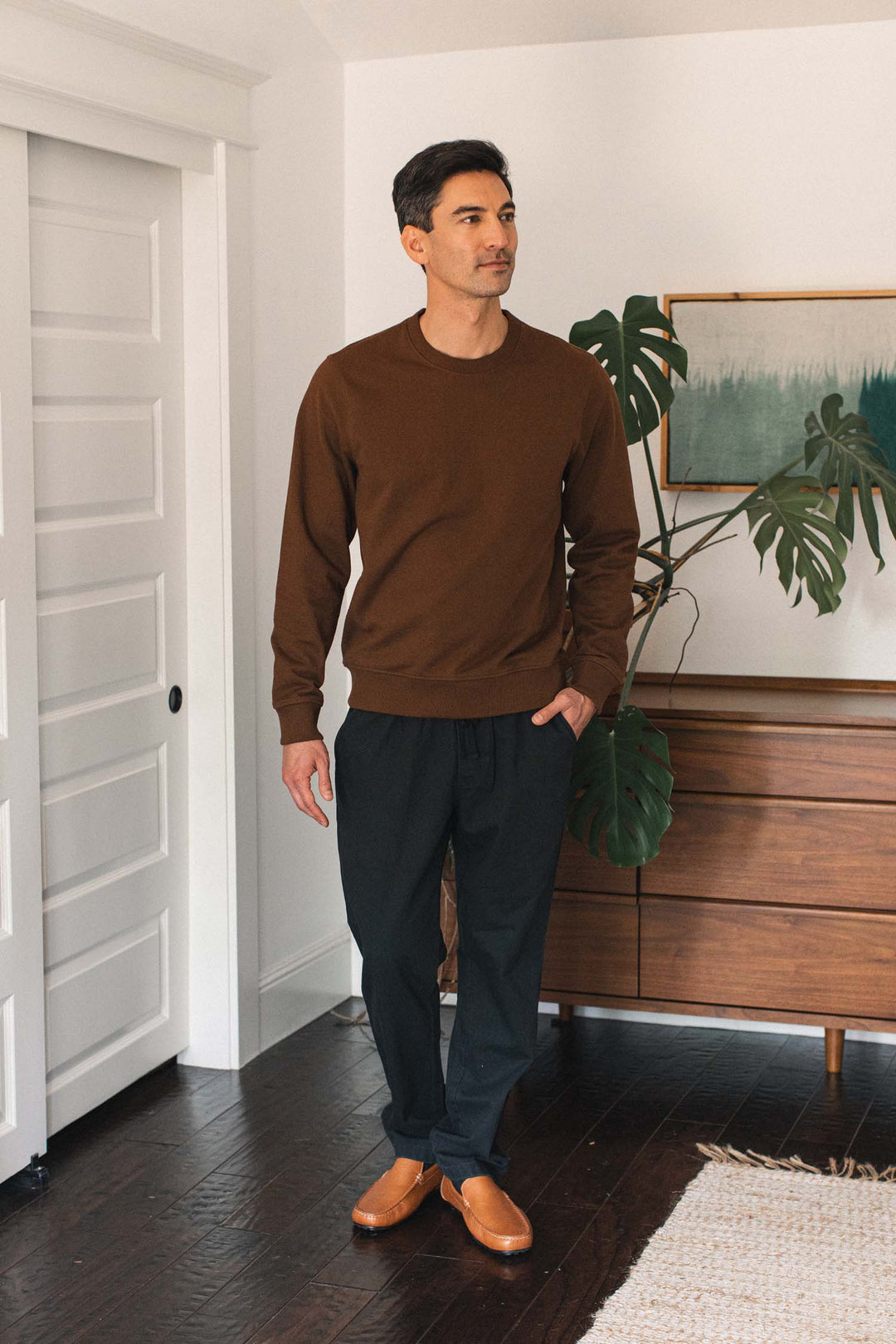 A man stands indoors near a wooden dresser and plant, wearing the Bridge & Burn Hayden Crew Sweatshirt in Cumin, paired with black pants and tan shoes. The room features white walls, a closed white door, and a framed abstract artwork on the wall.