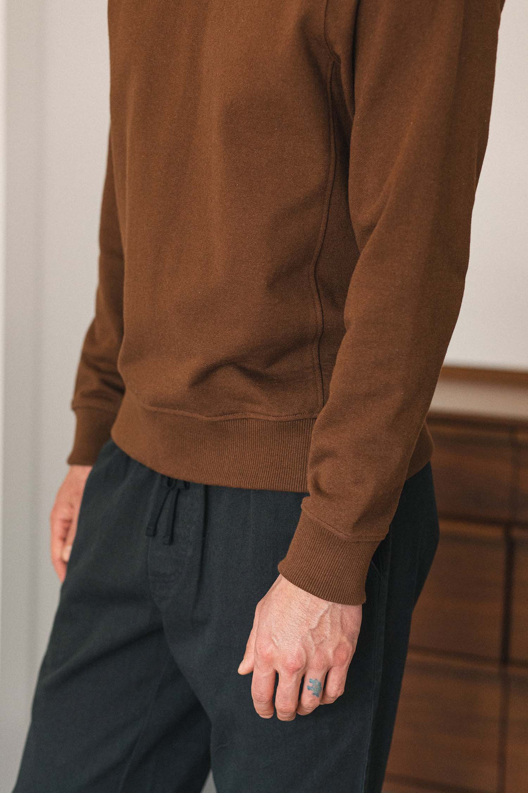 A person is shown indoors wearing the Bridge & Burn Hayden Crew Sweatshirt in Cumin with dark pants, their arm relaxed by their side. The image captures only the torso and upper legs, with a wooden dresser visible in the background.
