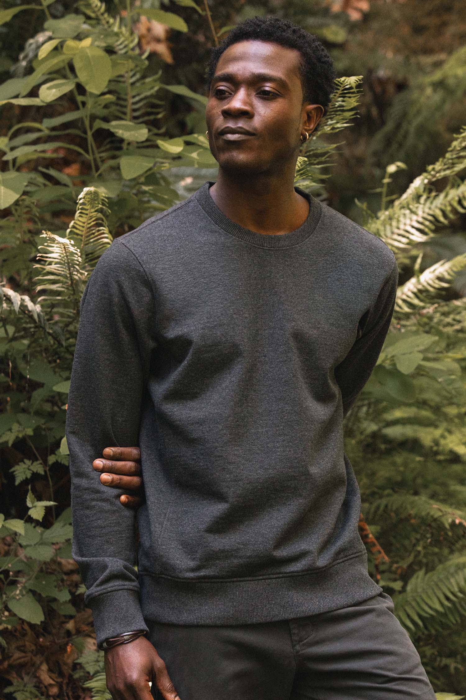 A person wearing the Bridge & Burn Hayden Crew Sweatshirt in Graphite stands outdoors among green fern plants, looking to the side with a calm expression.